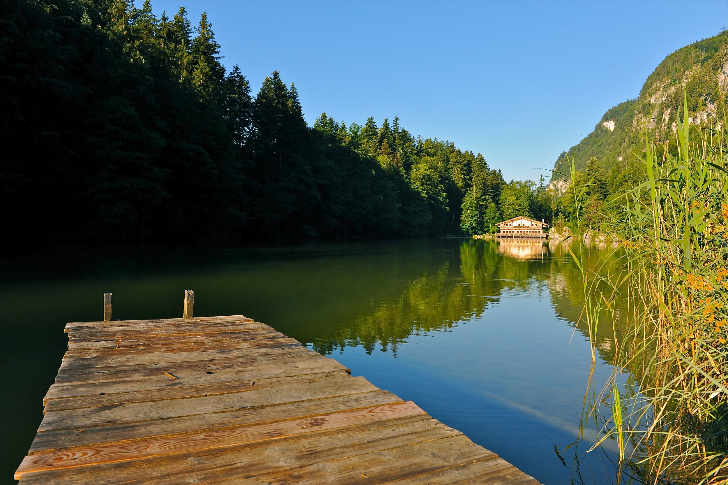 Holzsteg am Ufer eines ruhigen Sees, umgeben von grünen Bäumen und Bergen, mit einem Haus am Wasser im Hintergrund, bei sonnigem Himmel