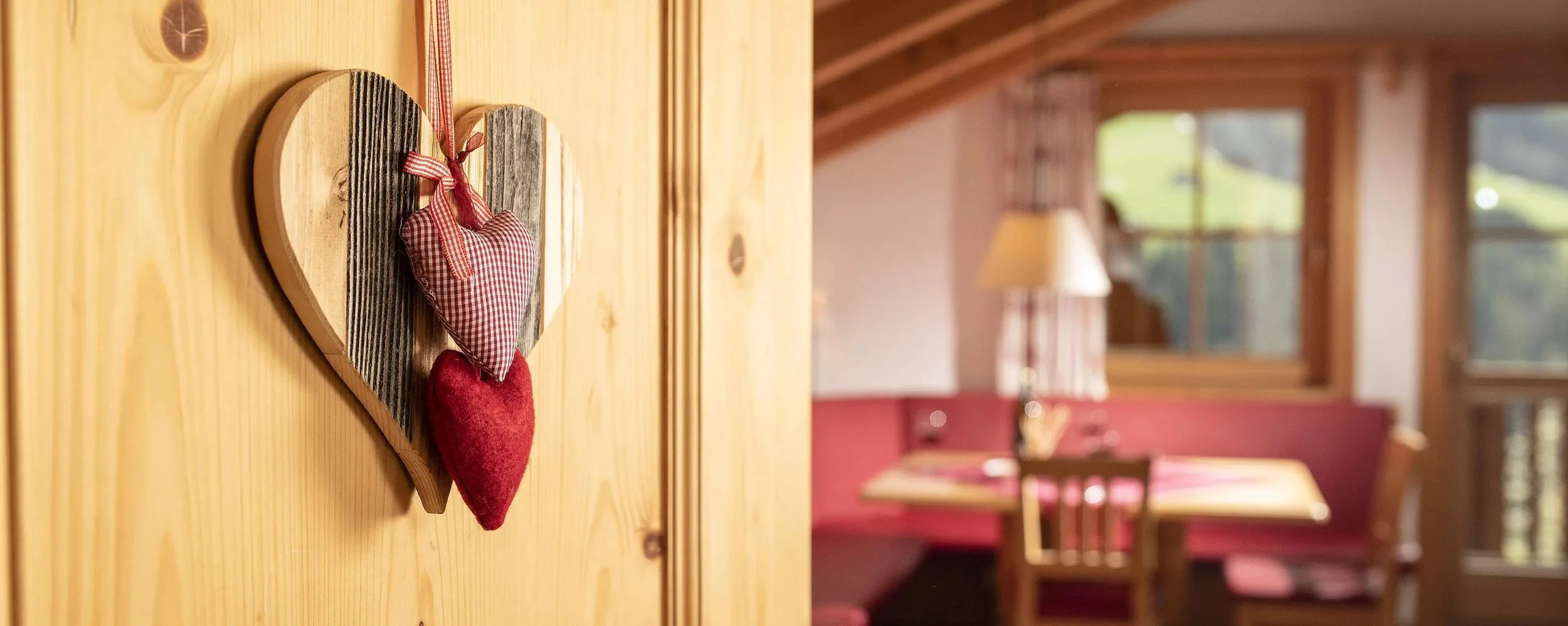Ein Herz aus Holz mit zwei Stoffherzen, ein rotes Filzherz und ein kleines Stoffherz in rot-weißem Karo, hängen an einer Holztür in einem gemütlichen Raum mit Holzbalken und einem Esstisch im Hintergrund.