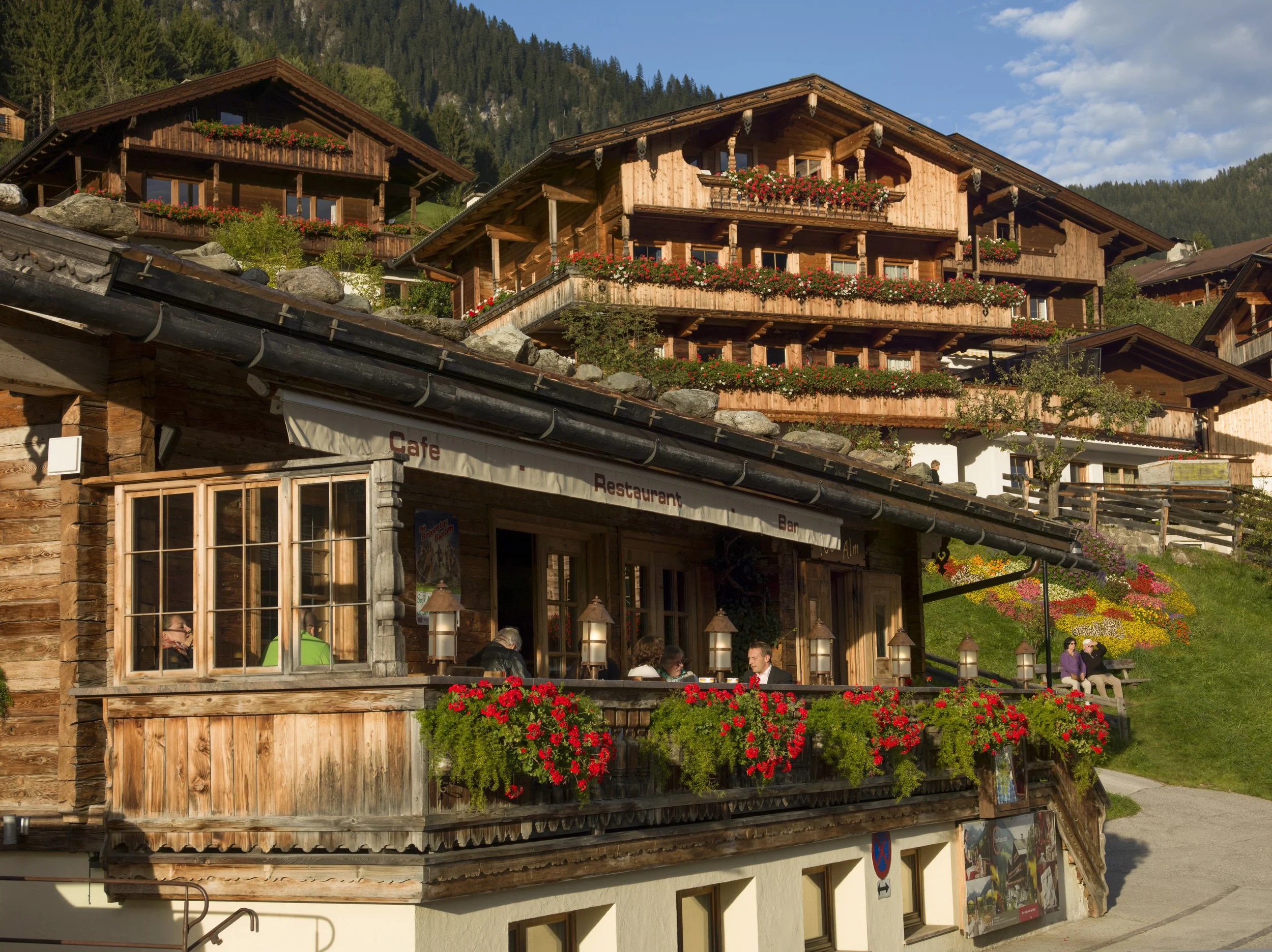 Traditionelles Chalet im alpinen Stil mit Holzfassade, Blumen am Balkon, inmitten grüner Berge, mit einem Café und Restaurant im Vordergrund, in dem Menschen sitzen und sich unterhalten.