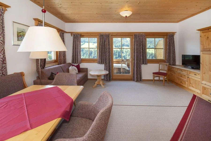 Gemütliches Wohnzimmer mit Holzwänden und -decken, Fenster mit Vorhängen, Sitzbereich mit Sofa und Stühlen, Esstisch mit roter Tischdecke, Fernseher in Holzschrank, Blick auf bergiges Außenlayout