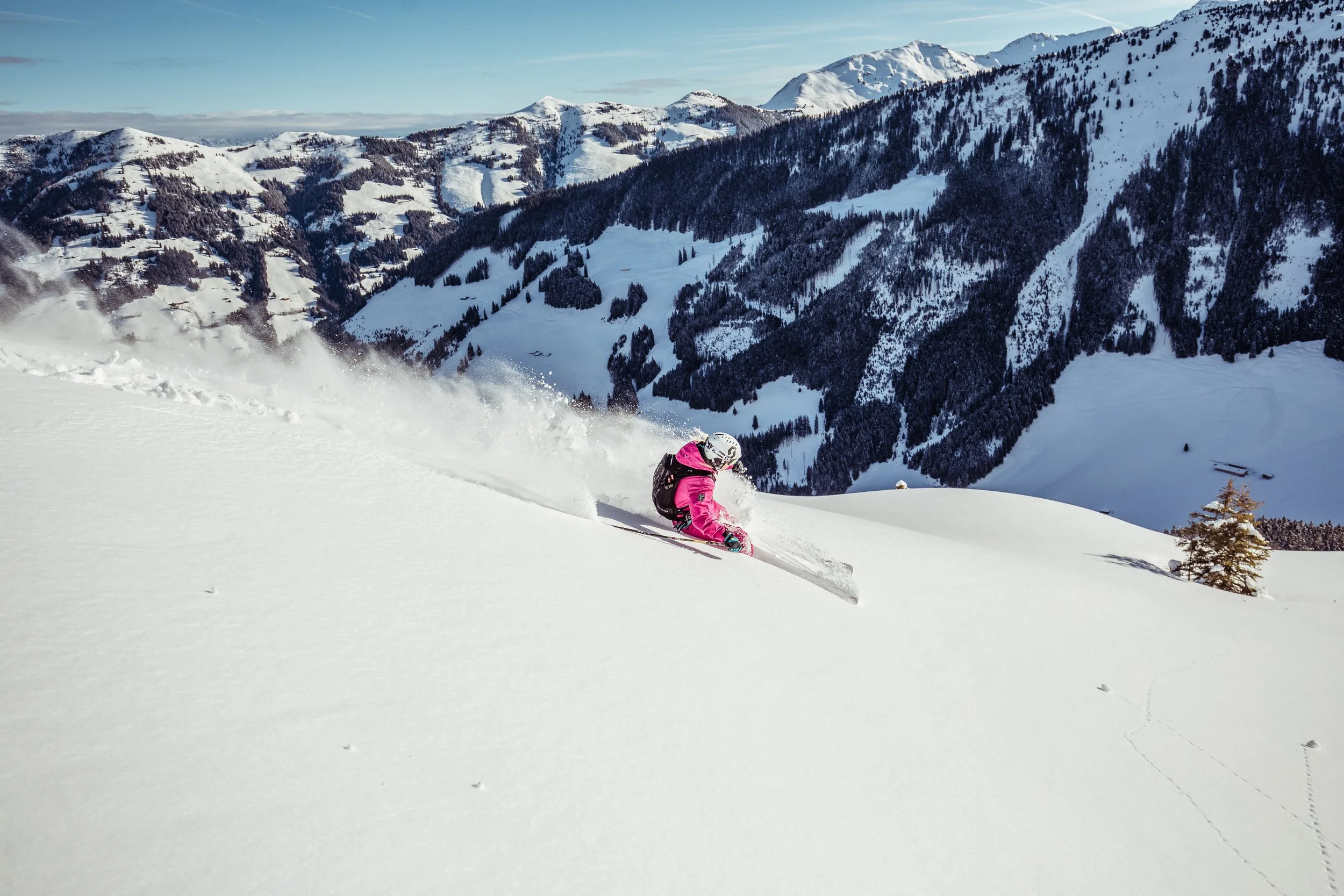 Skifahrer in pinke Winterkleidung fährt den Hang hinunter in einer verschneiten Berglandschaft mit Bergen und Bäumen im Hintergrund.