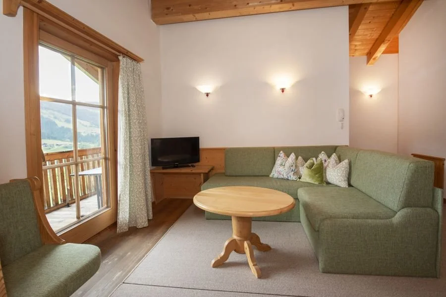 Ein gemütliches Wohnzimmer mit einem grünen Sofa, einem kleinen runden Holztisch, einem Fernseher, einer Balkon-Tür und Wandlampen.