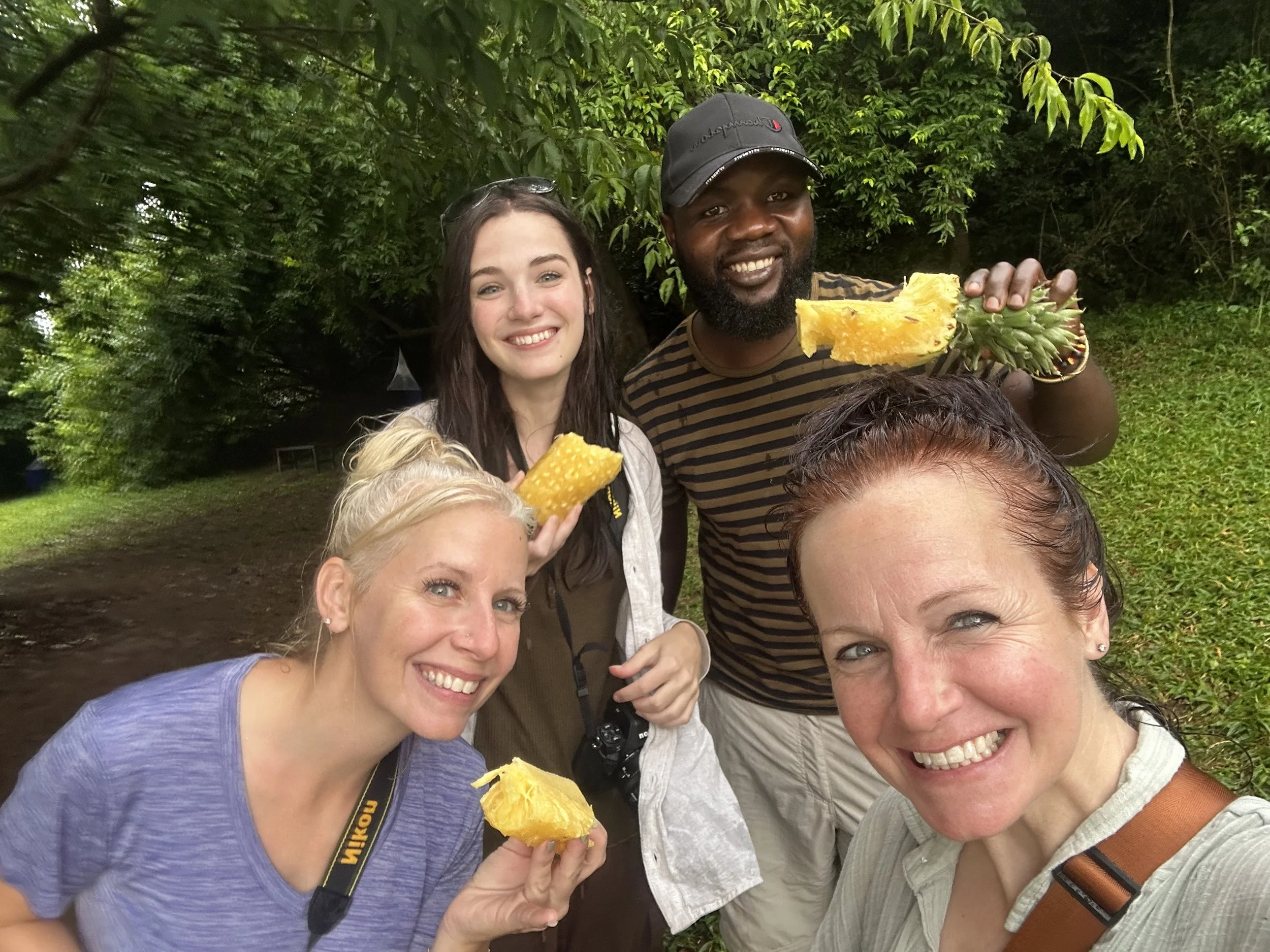 A group of five smiling people outdoors, holding slices of pineapple, with green trees and foliage in the background.