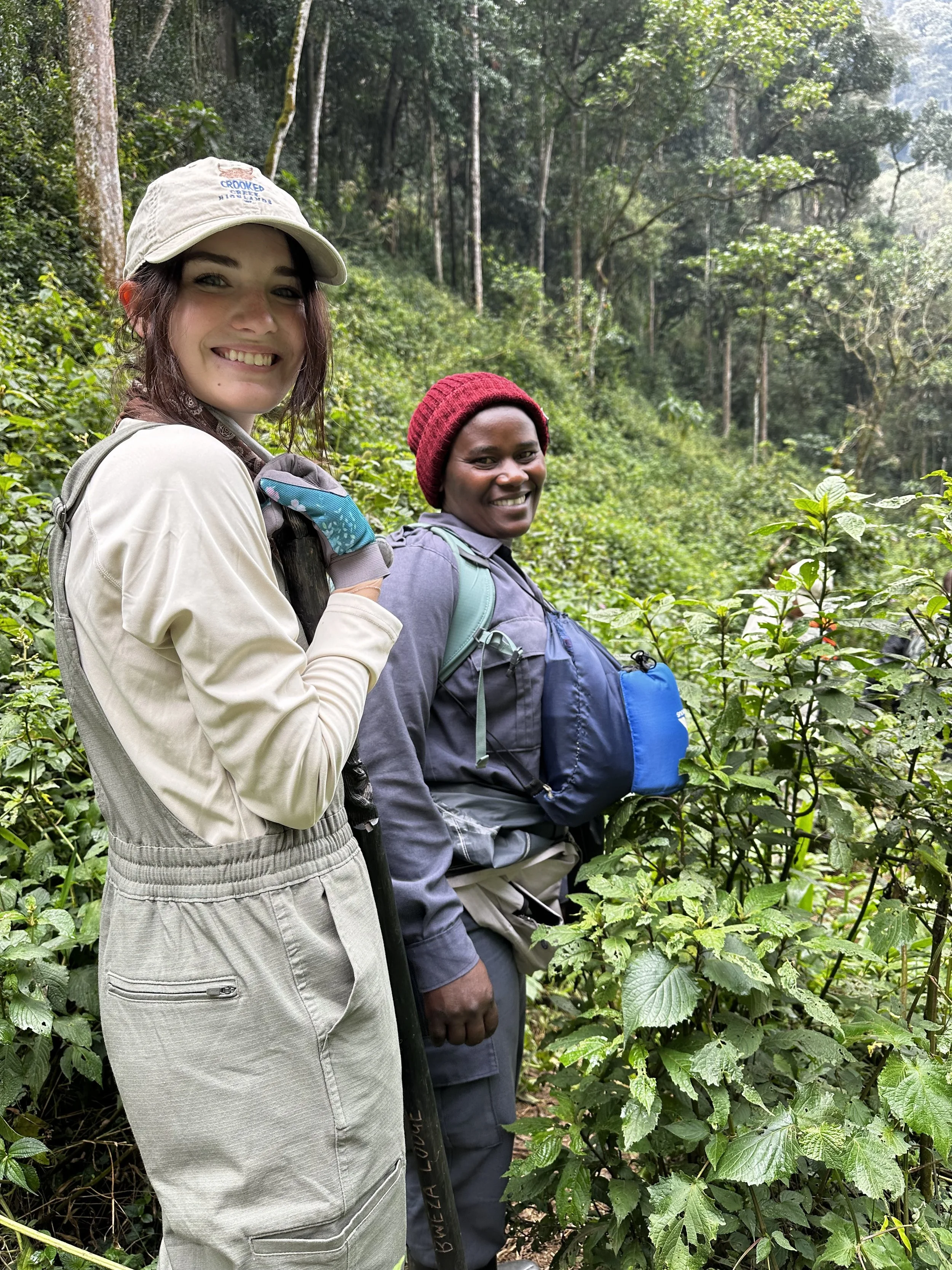 Two women in outdoor clothing and backpacks stand in a lush green forest surrounded by dense trees and plants, smiling at the camera.