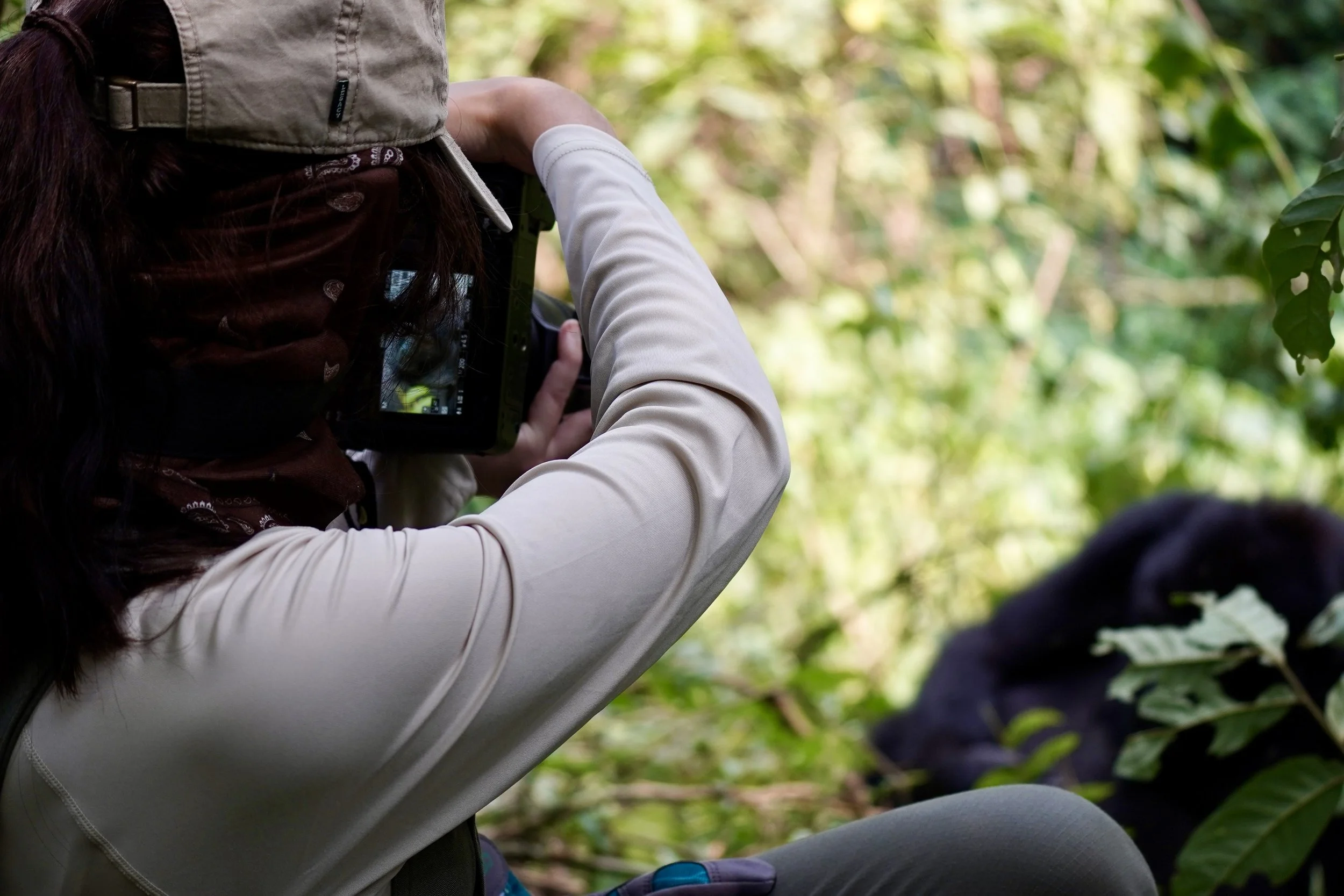 A person wearing a beige hat and white long-sleeve shirt looking through a camera in a green, outdoor setting with blurred foliage in the background.