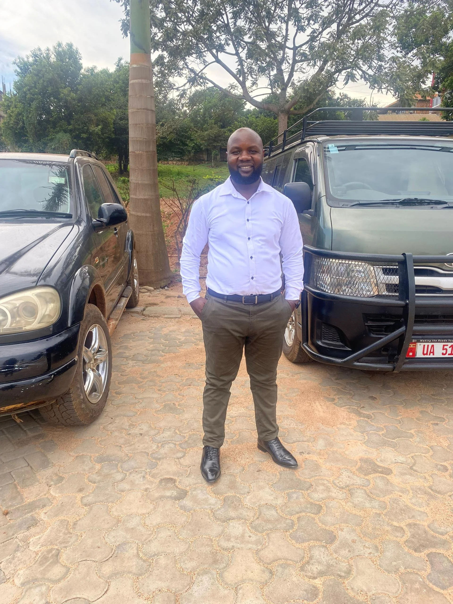 A man standing outdoors on a paved surface between two parked vehicles with greenery and trees in the background, smiling at the camera.