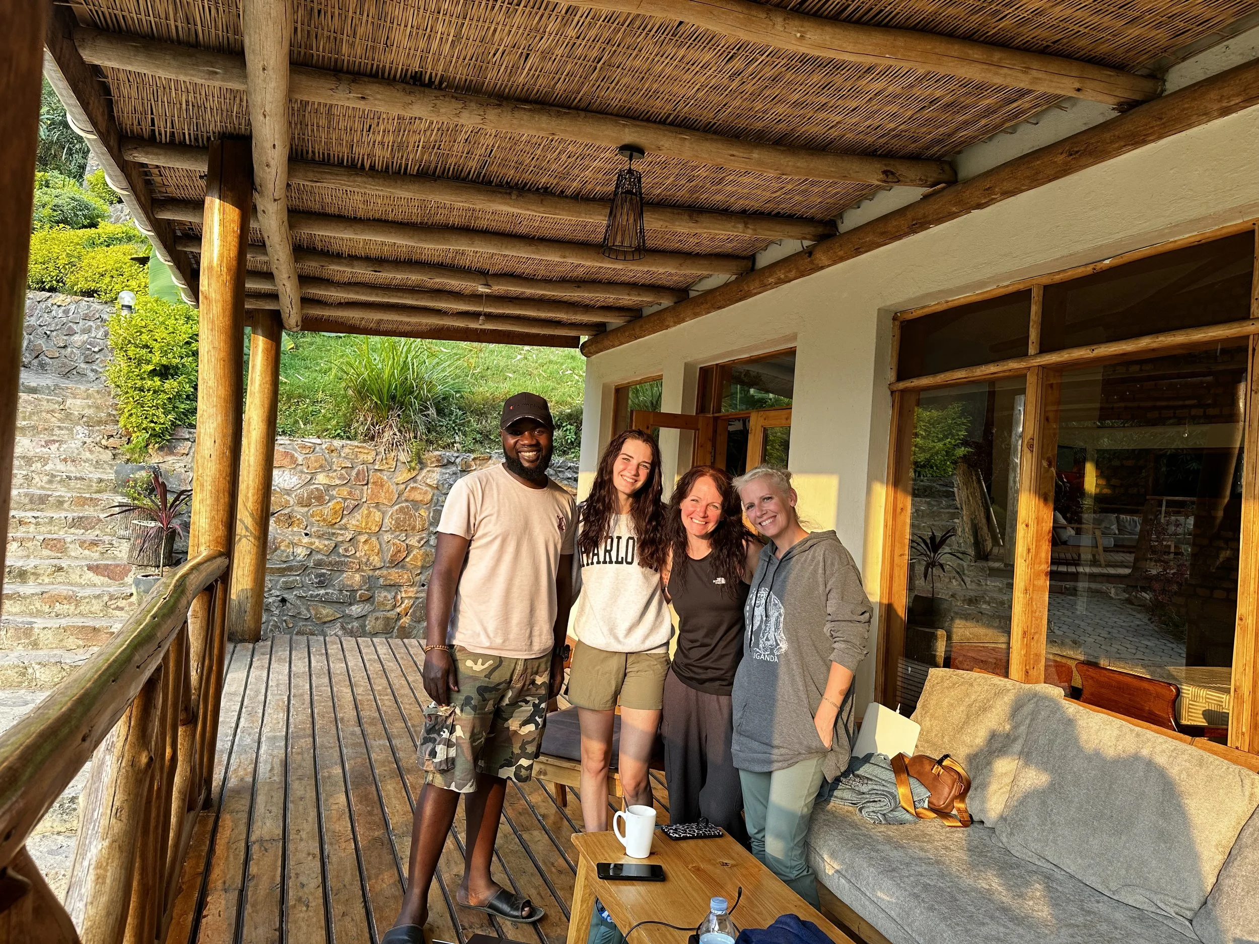 A group of five people standing on a wooden porch, smiling and posing for the photo. They are in front of a house with large windows and surrounded by greenery, with a stone retaining wall and plants in the background. The porch has a bamboo ceiling and wooden railings.
