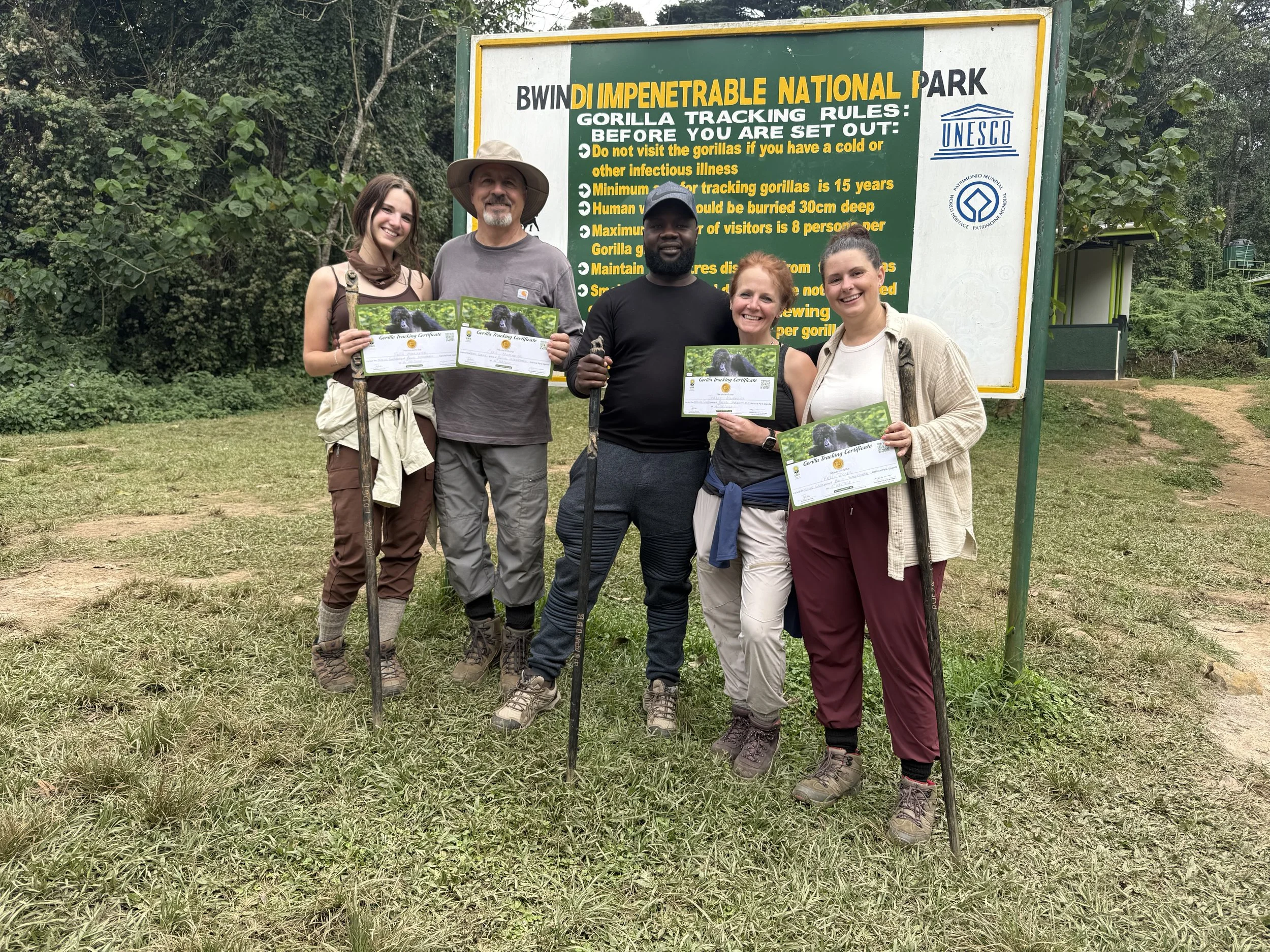 A diverse group of five people standing outdoors in front of a sign at Bwindi Impenetrable National Park, holding certificates and smiling. The sign provides rules for gorilla tracking in the park, which is a UNESCO World Heritage Site, surrounded by lush green foliage.