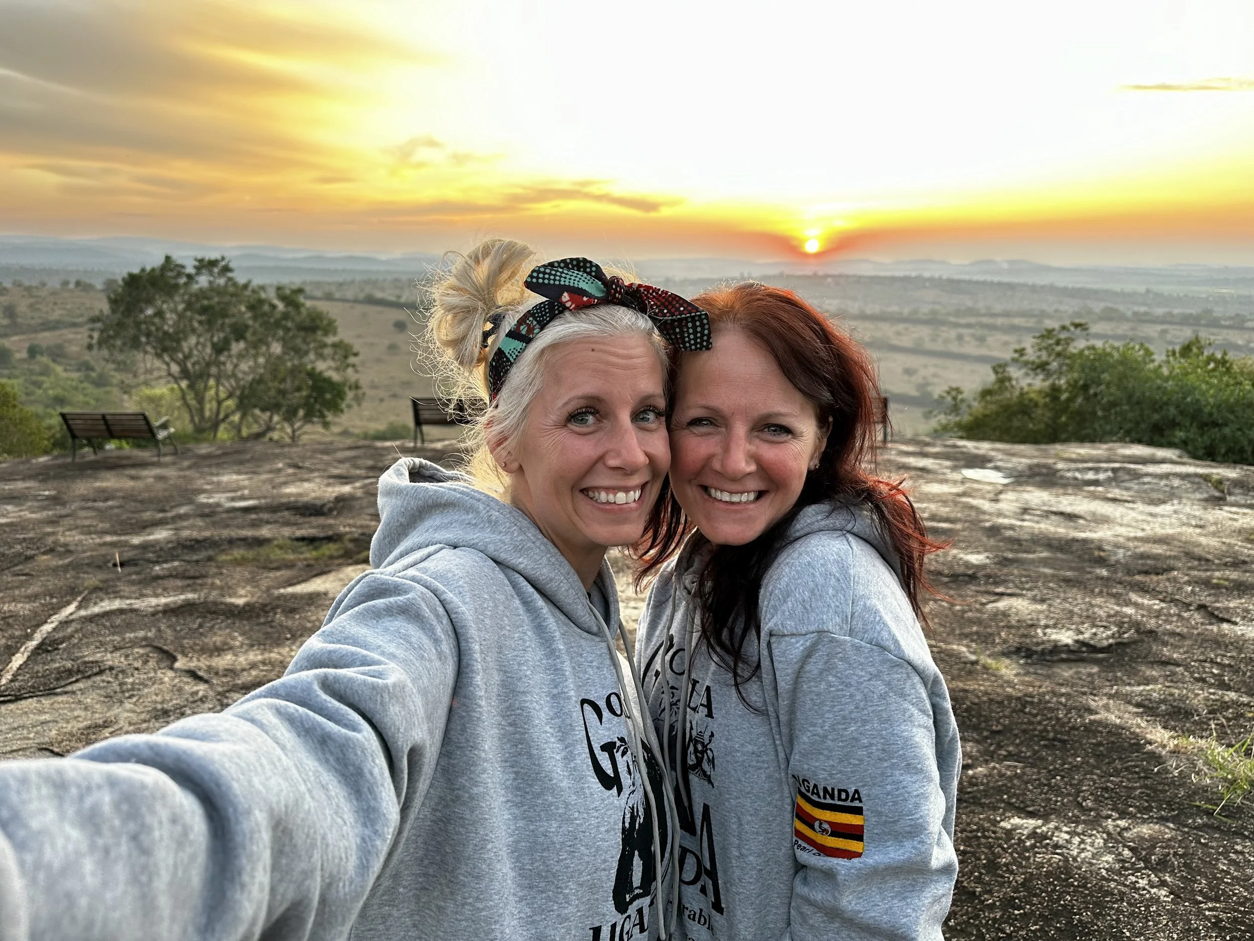 Two women taking a selfie outdoors at sunset, with a scenic landscape and benches in the background.