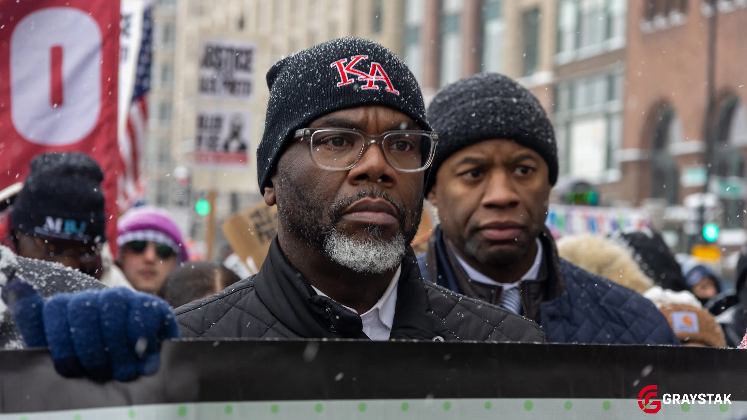 Two men wearing black beanies, one with glasses and a gray beard, and the other with a serious look, participating in a protest or rally in a city street during winter with snow falling and a crowd of people and buildings in the background.