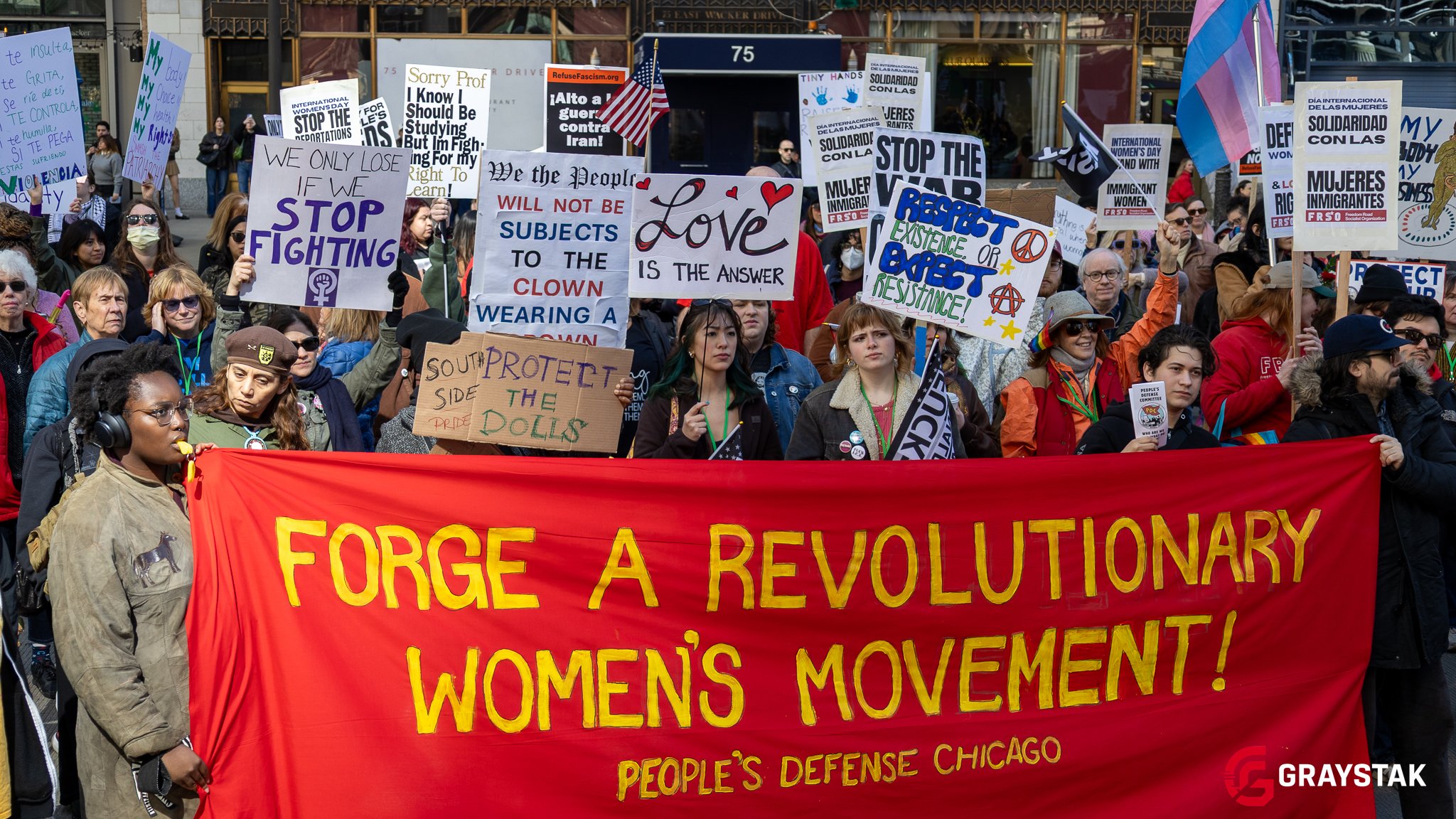 A large crowd of diverse protesters holding various signs and banners during a demonstration, with a prominent red banner in the foreground reading 'FORGE A REVOLUTIONARY WOMEN'S MOVEMENT!' in yellow text.