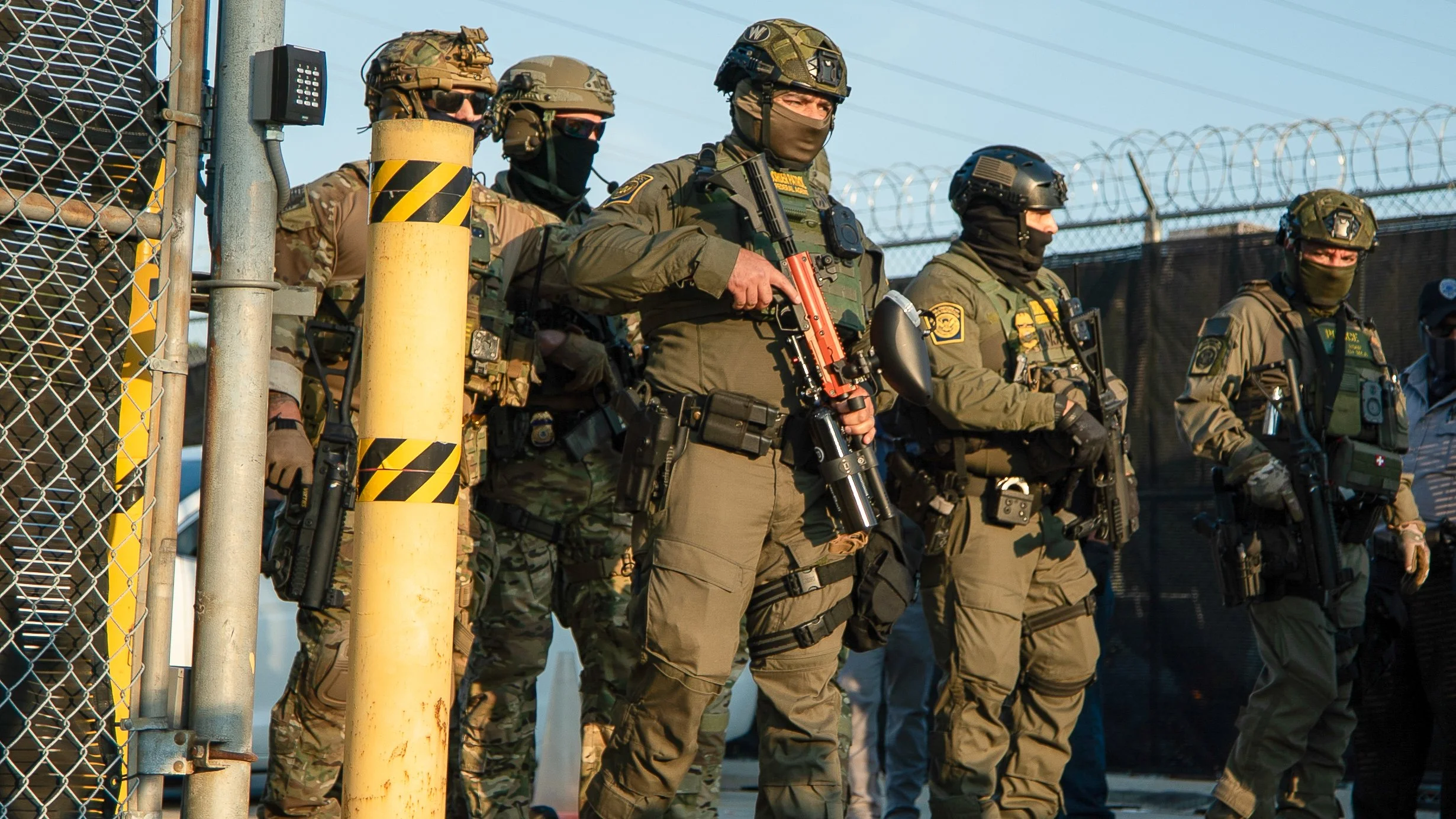 Military personnel standing in a line behind a chain-link fence, wearing tactical gear, helmets, and masks.