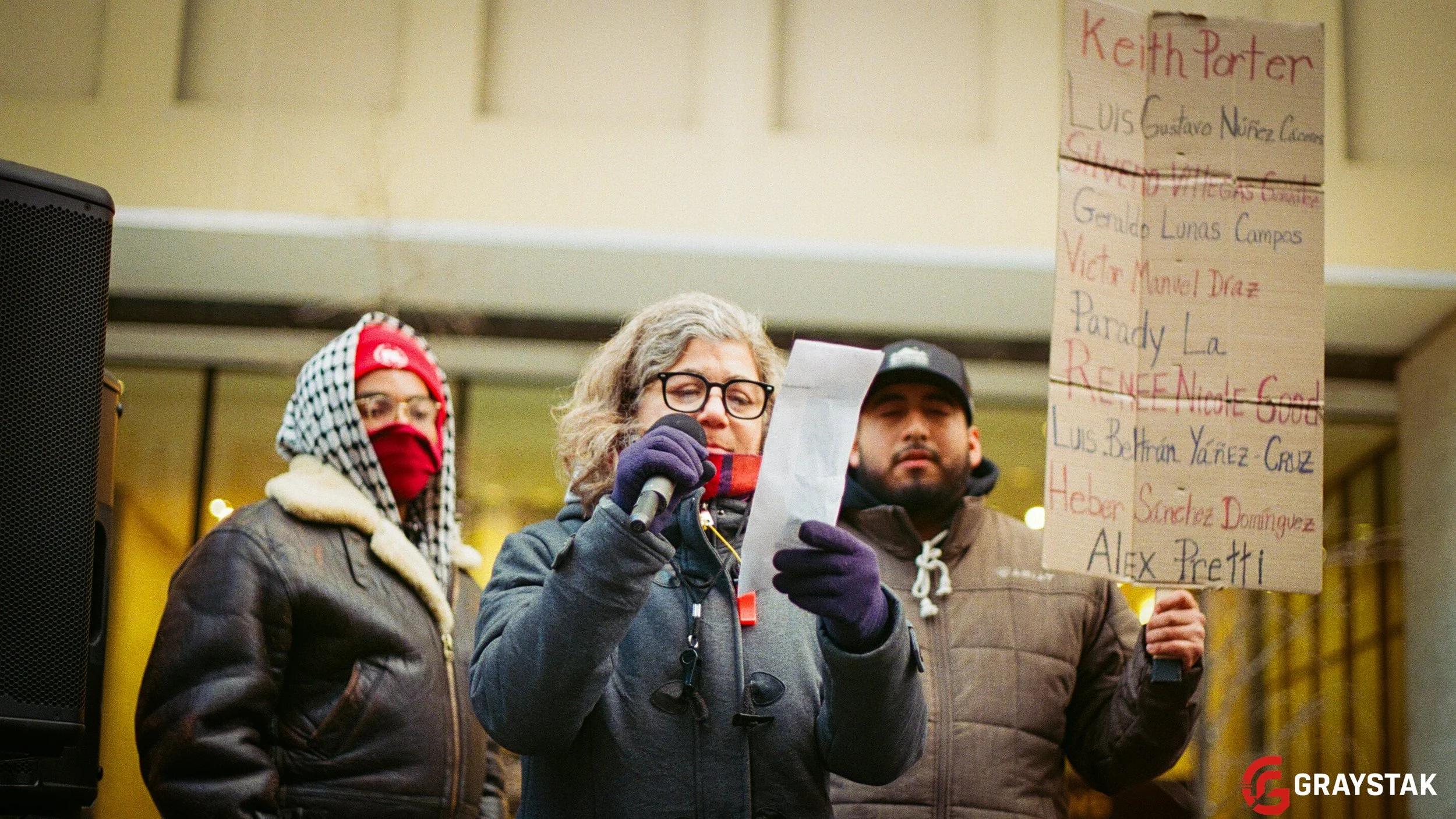 A group of people participating in a protest or demonstration indoors. One woman is speaking into a microphone, holding a piece of paper, and wearing glasses, a mask, and gloves. Behind her, a man is holding a large sign with text, and other individu