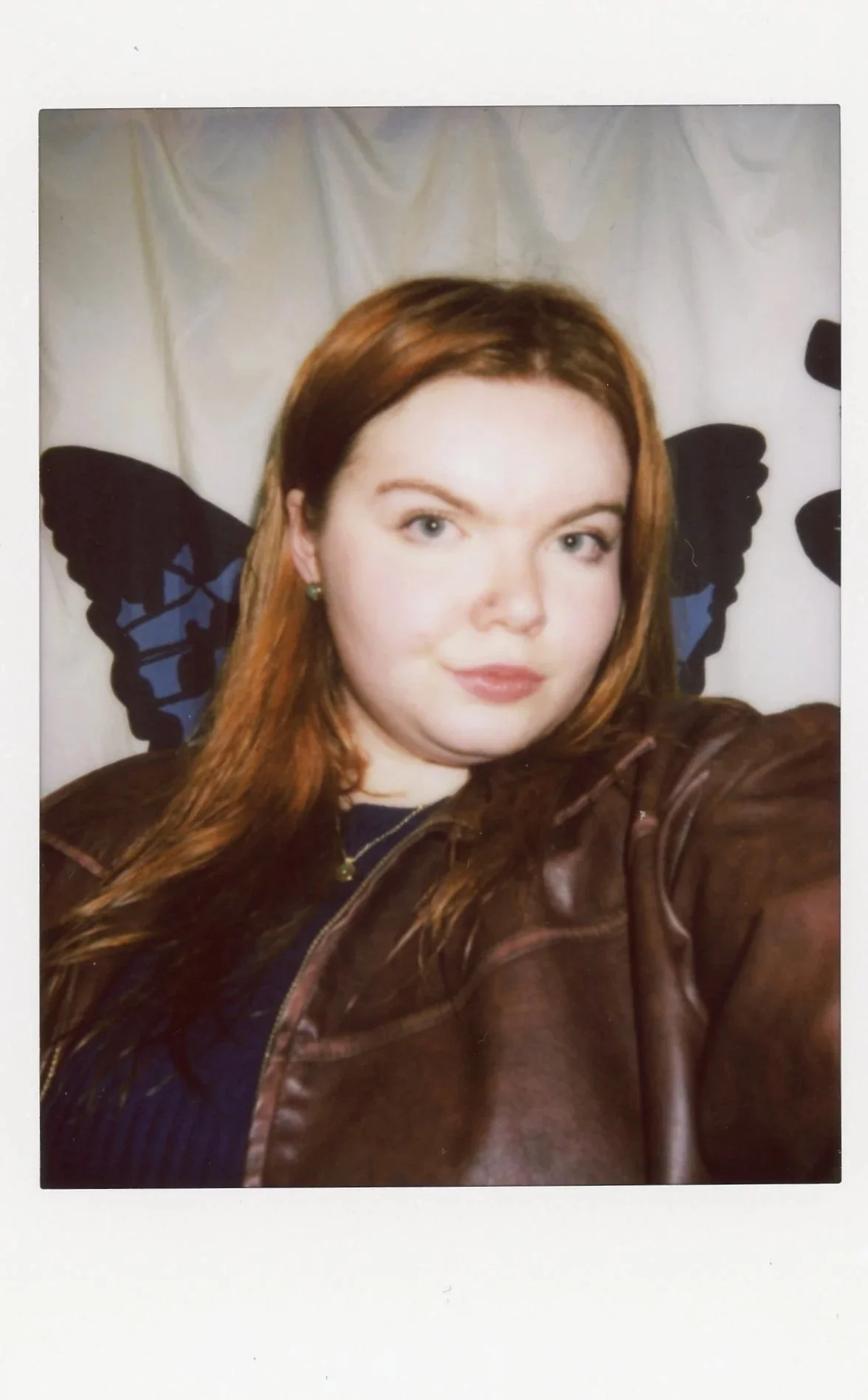 A young woman with long red hair taking a selfie. She is wearing a brown leather jacket and has a butterfly-shaped hair clip. There is a butterfly-shaped wall decoration behind her.