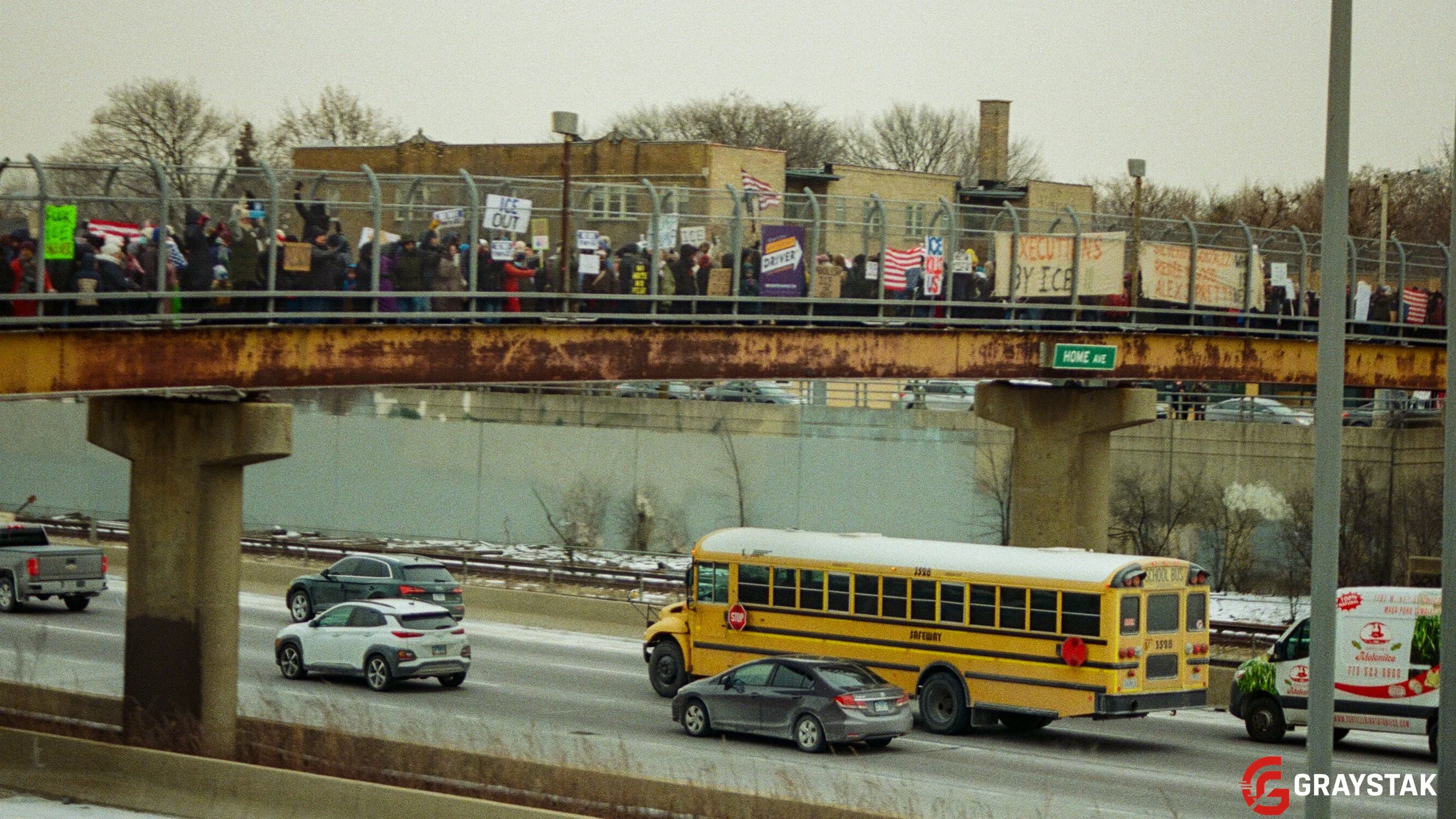 A group of people are gathered on a bridge holding signs and flags, protesting or demonstrating, with trees and buildings in the background.