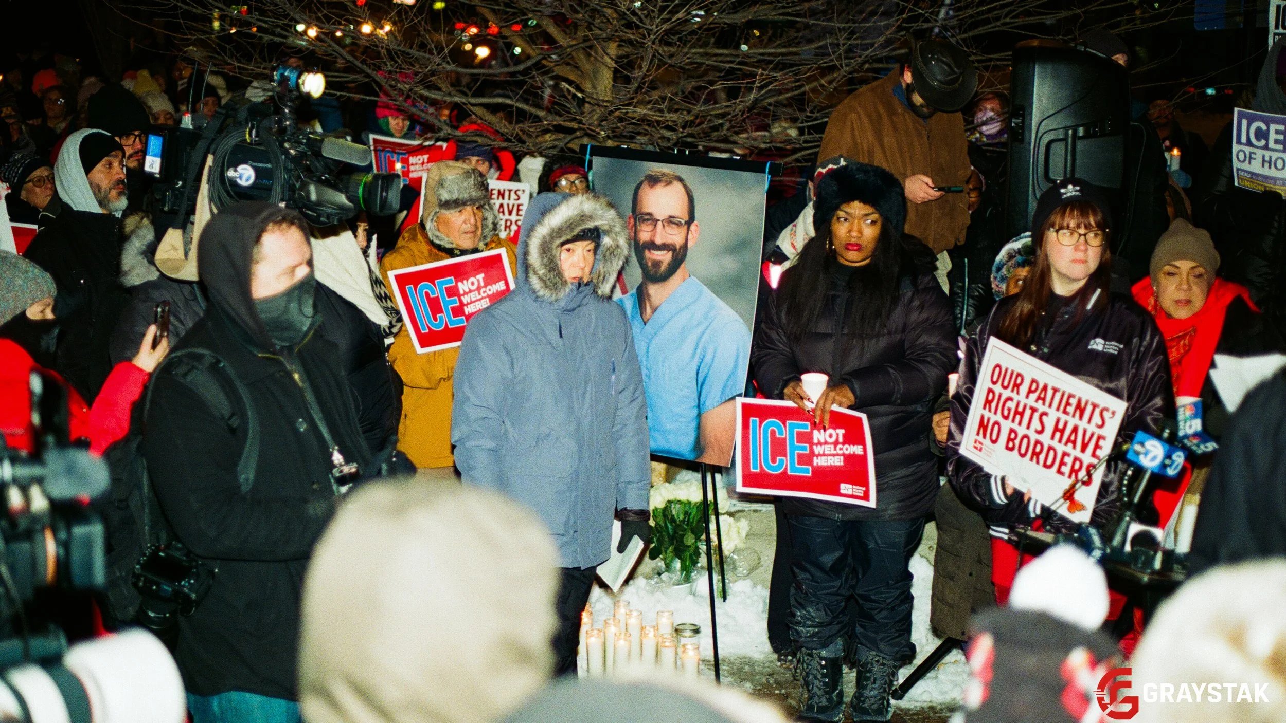 A group of people holding protest signs at an outdoor event during winter. Signs include messages like "ICE NOT WELCOME HERE" and "OUR PATIENTS' RIGHTS HAVE NO BORDERS." Some people are wearing warm clothing and there are candles on the ground.