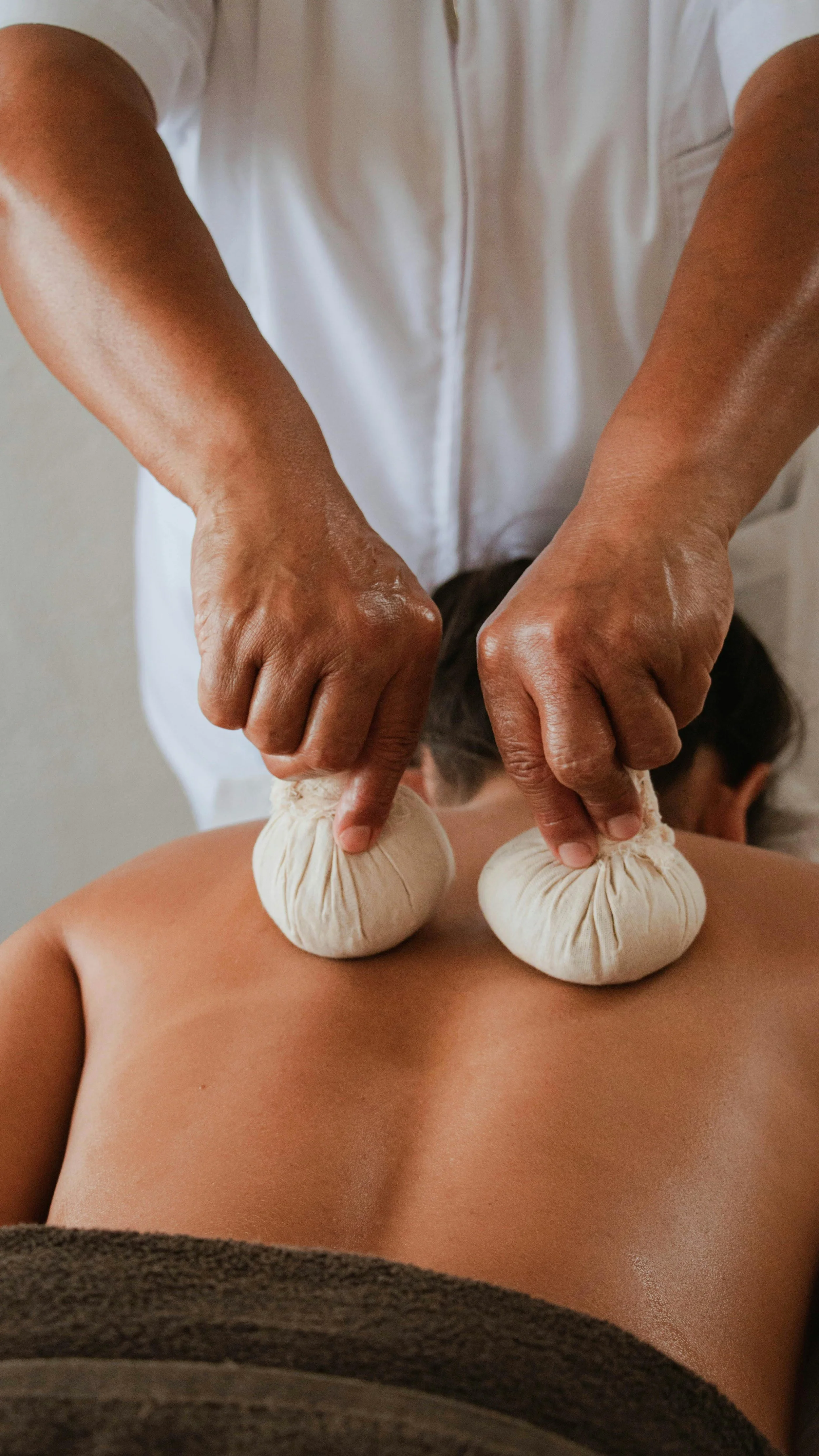A person receiving a hot stone massage with heated herbal compress balls on their back, while a massage therapist presses on the client's back.