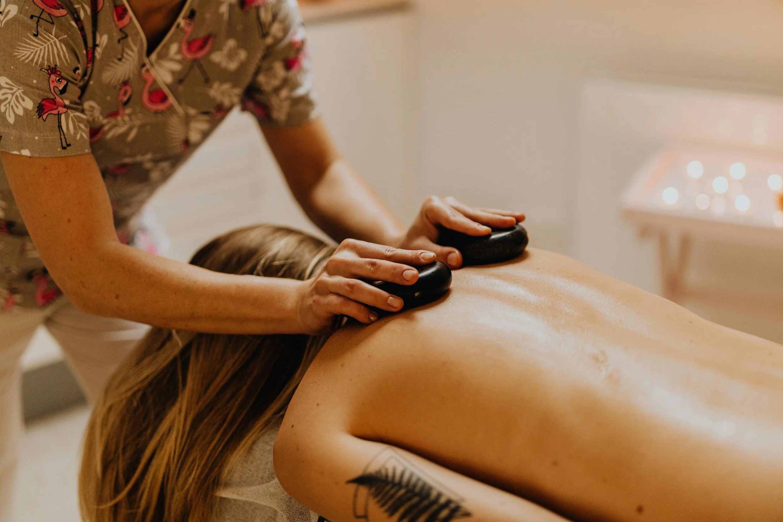 A person receiving a hot stone massage on their back, with the masseuse's hands placing smooth black stones on the person's back in a spa or massage room.