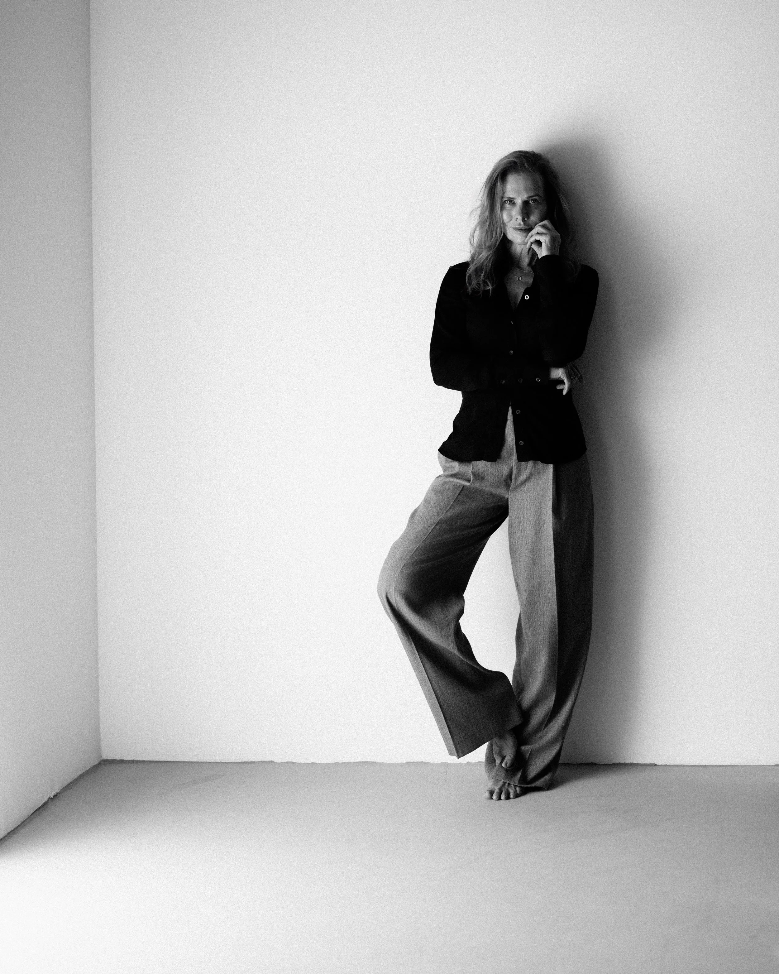 A woman standing barefoot in a corner, resting her back against the wall, with her hand near her face, wearing a dark blouse and loose-fitting pants, in black and white photograph.