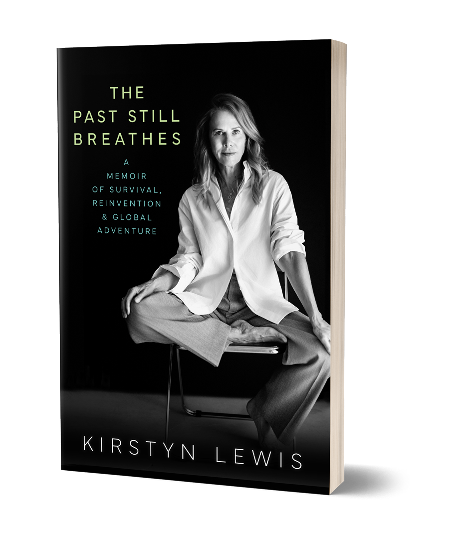 Black and white photo of a woman sitting cross-legged on a chair, wearing a loose white shirt and grey pants, in front of a black background, on the cover of a book titled "The Past Still Breaths" by Kirstyn Lewis.