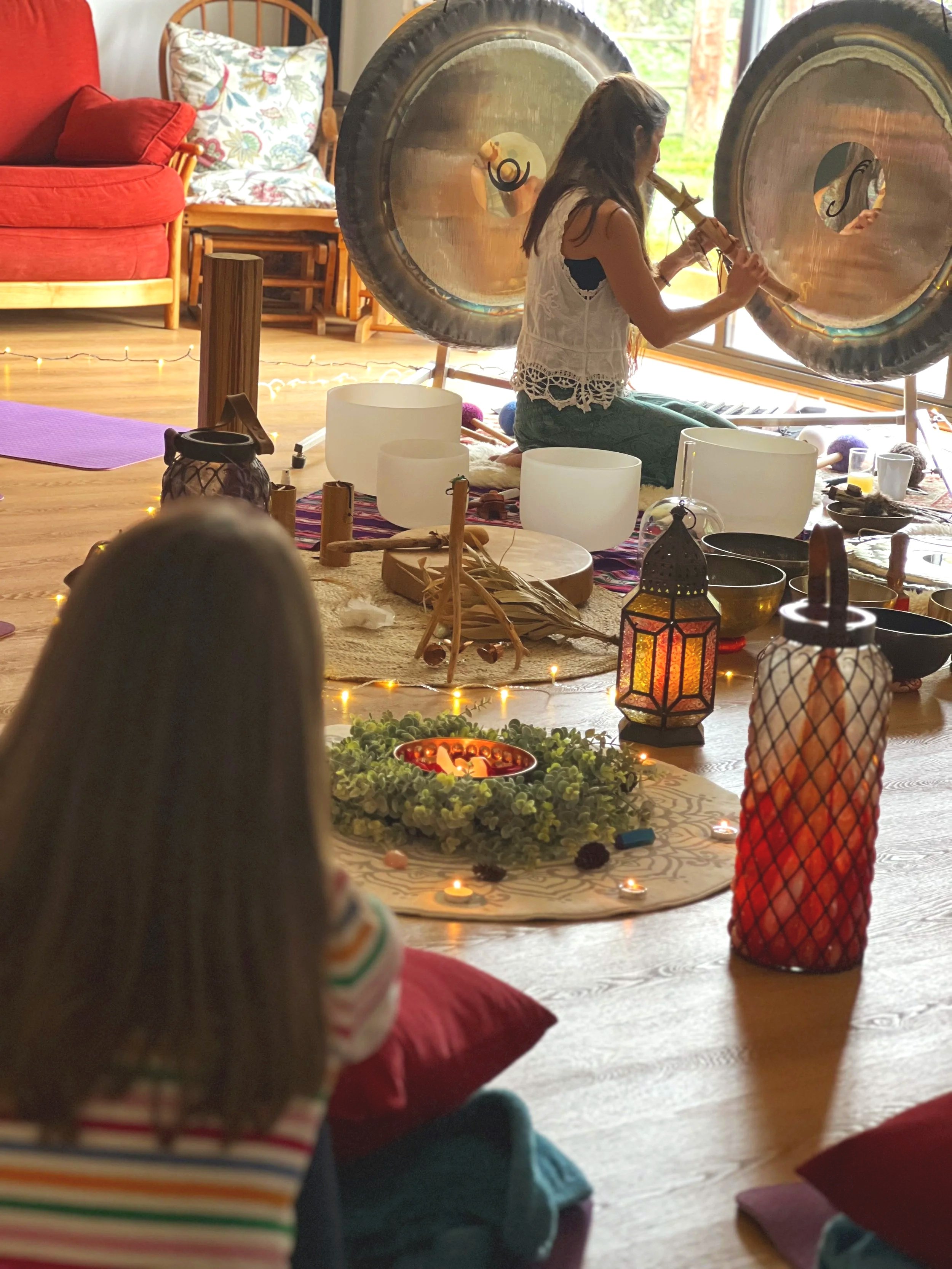 A woman playing a wind instrument in a room with large gongs, surrounded by candles and decorative objects, pets, and children sitting on the floor.