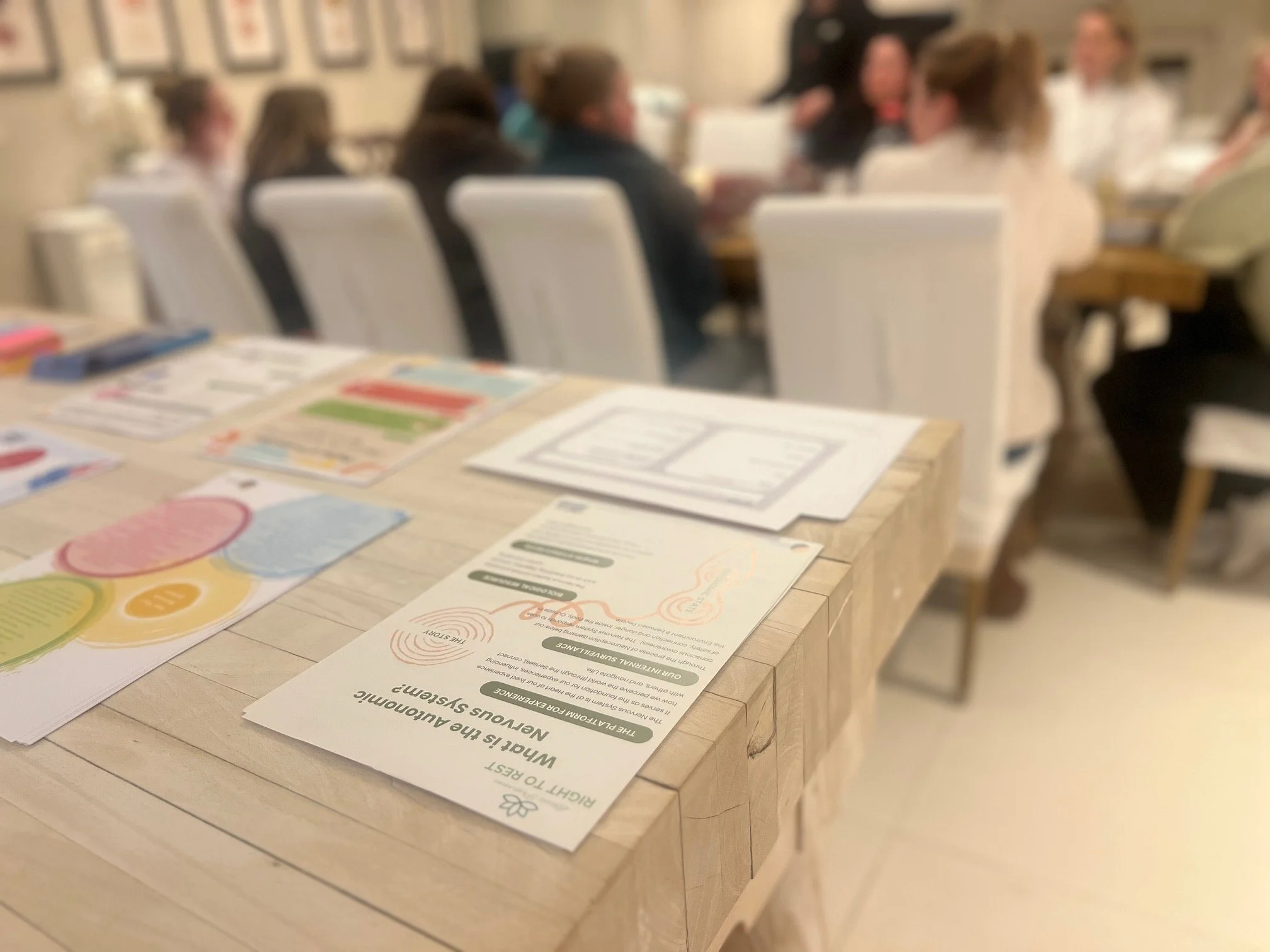A blurred photo of a meeting room with people sitting around a table. The foreground shows a wooden table with colorful informational flyers and a sheet of paper.