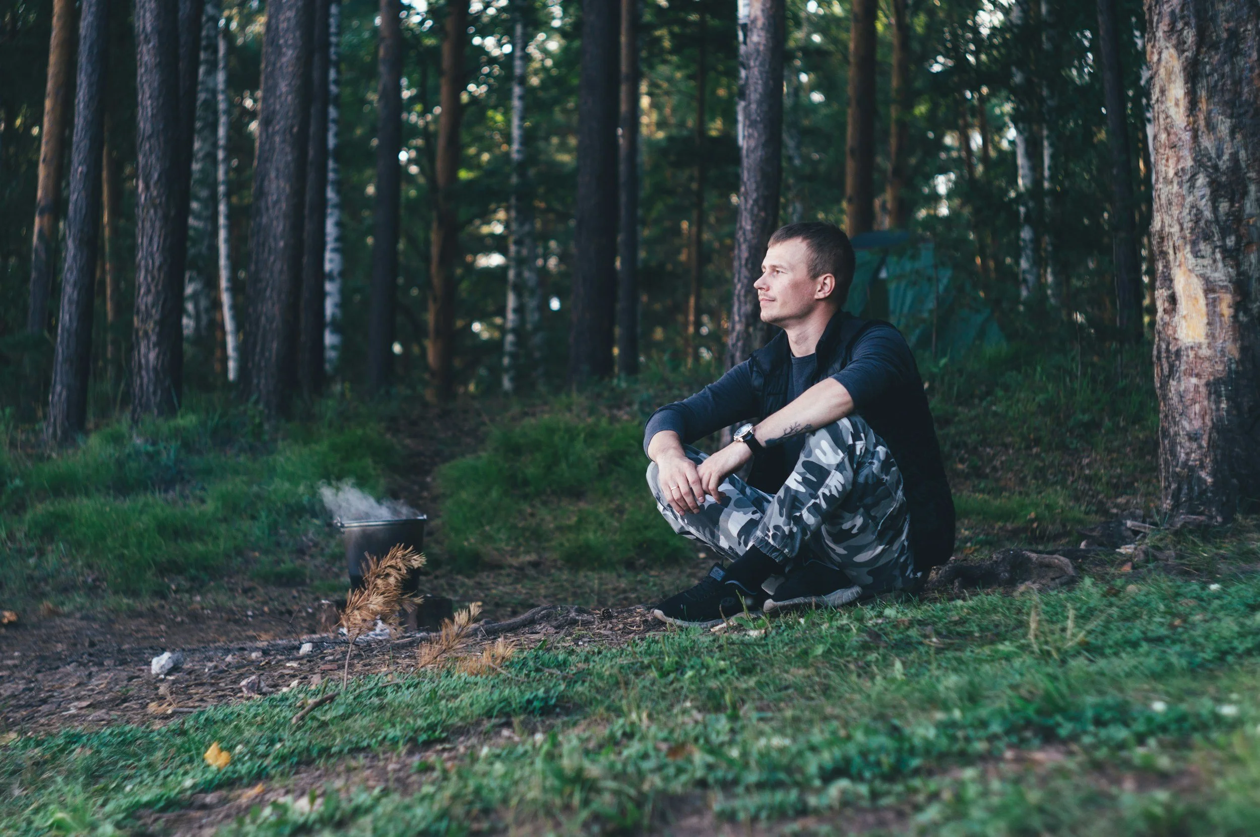 A man sitting on the grass in a forest, looking to the right, with a charcoal grill and a small grill brush nearby, surrounded by tall trees