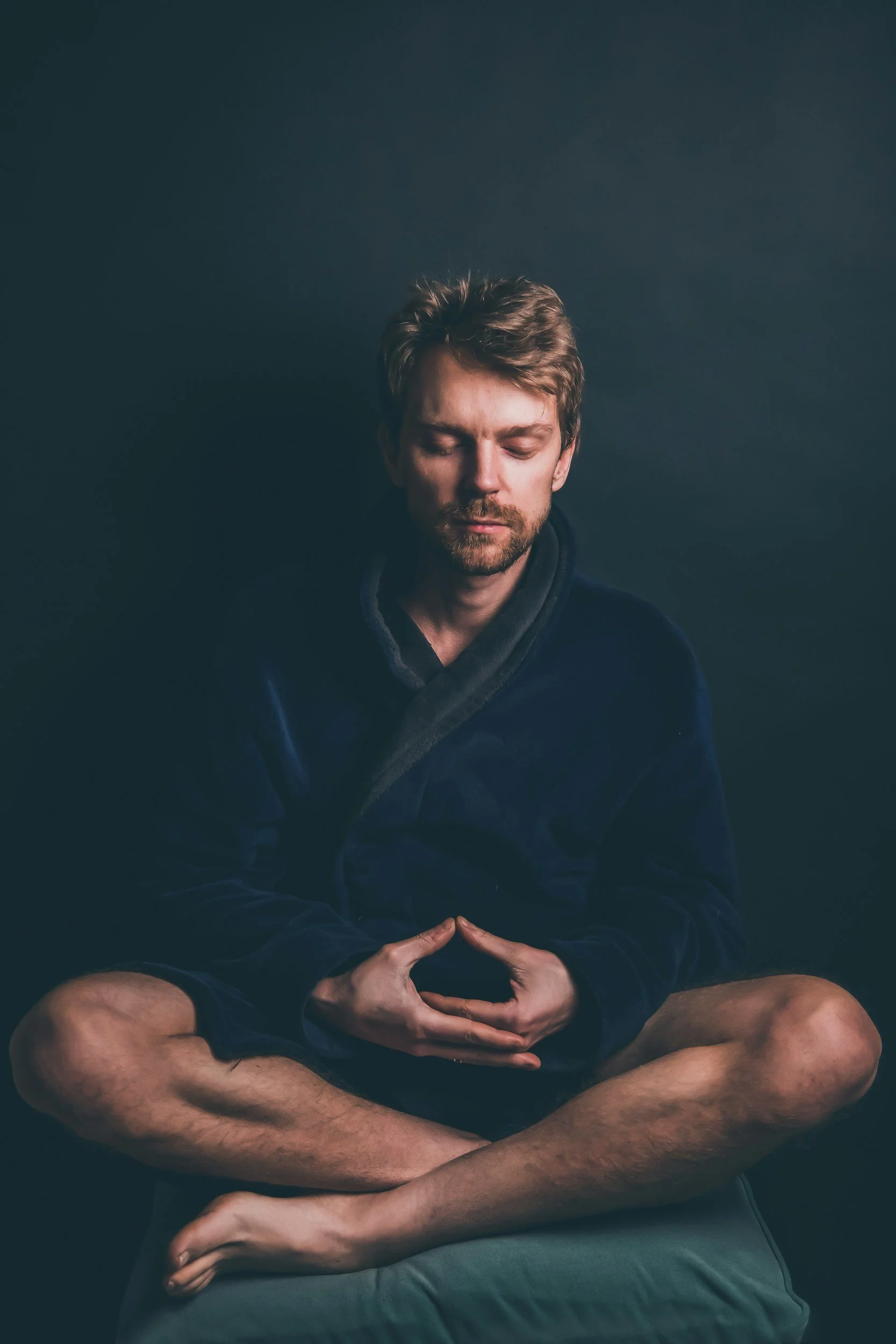 A man with closed eyes sitting cross-legged with hands resting in a meditative pose, against a dark background.