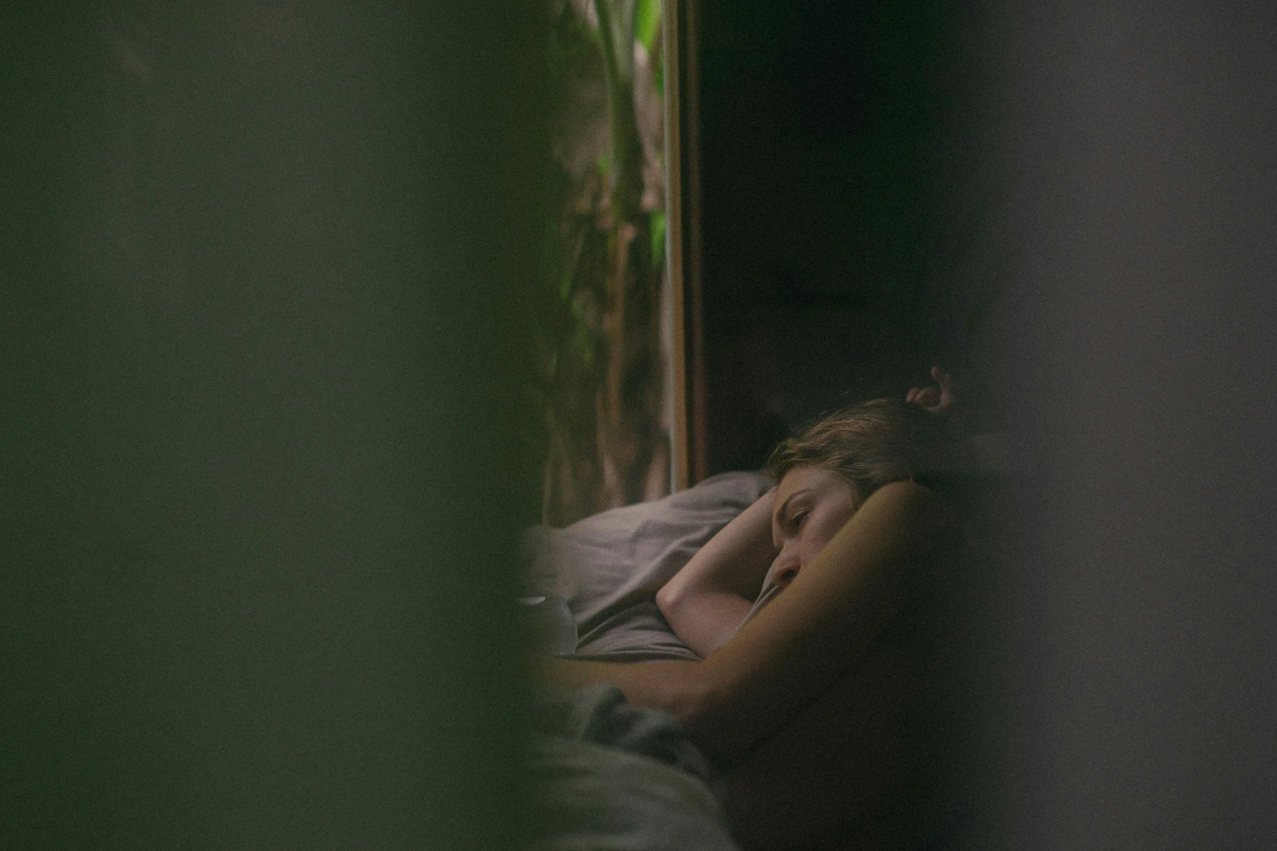 A person lying on a bed with hands behind their head, resting, next to a window with a view of greenery outside, at nighttime.