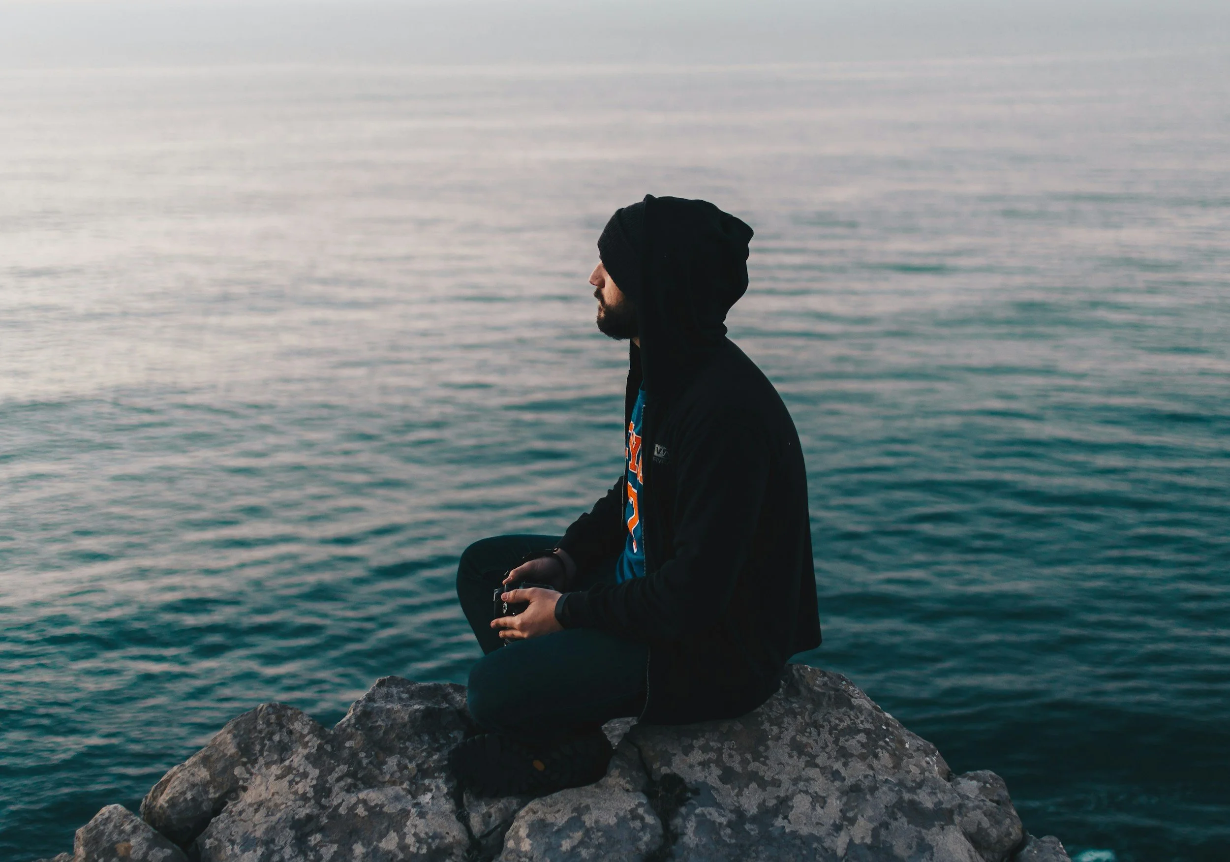 A person sitting on rocks near the water, wearing a black hoodie and looking out at the ocean.