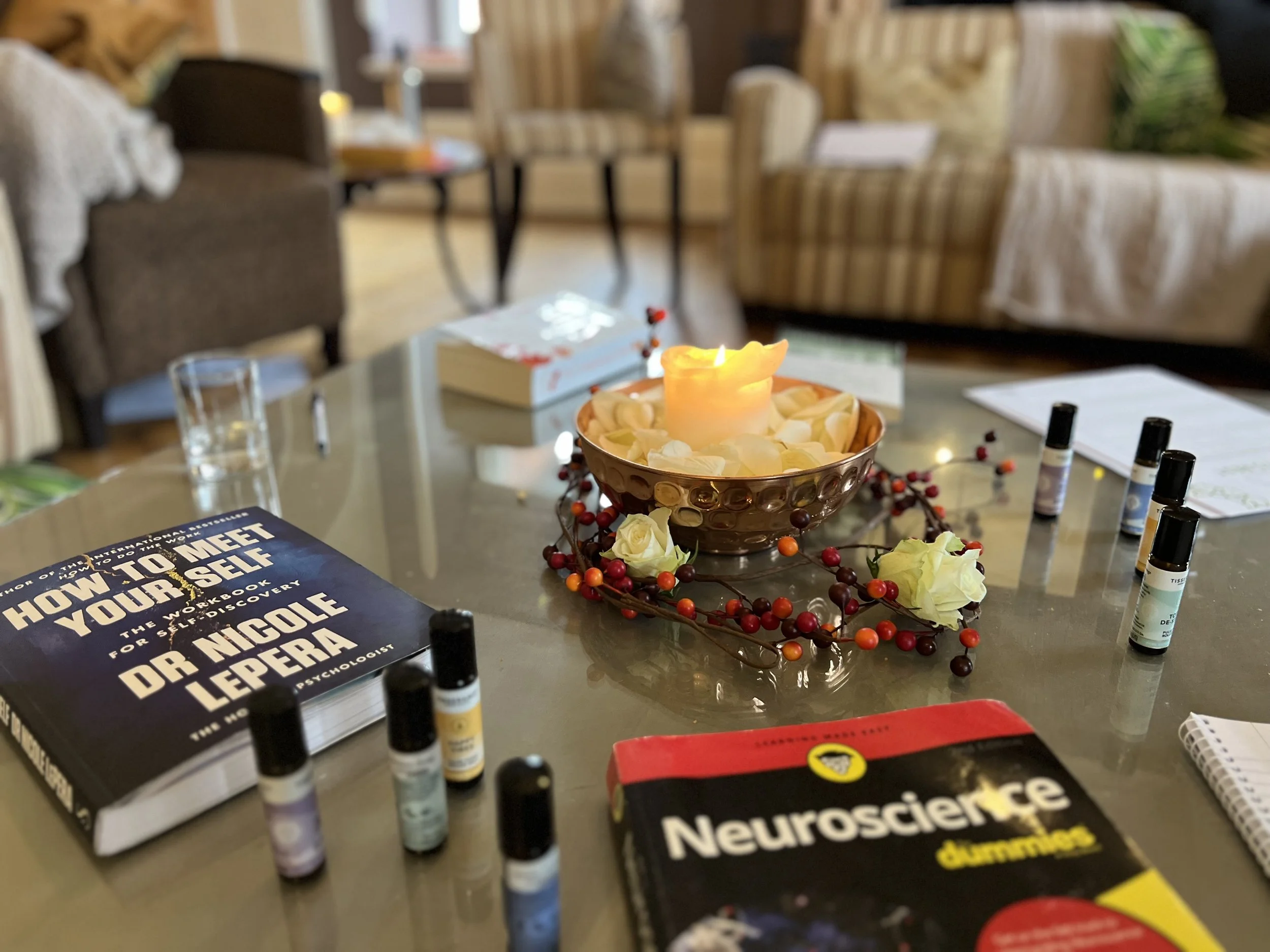 A table with books, pens, a candle, and decorative berries, in a cozy room with chairs and a patterned sofa in the background.
