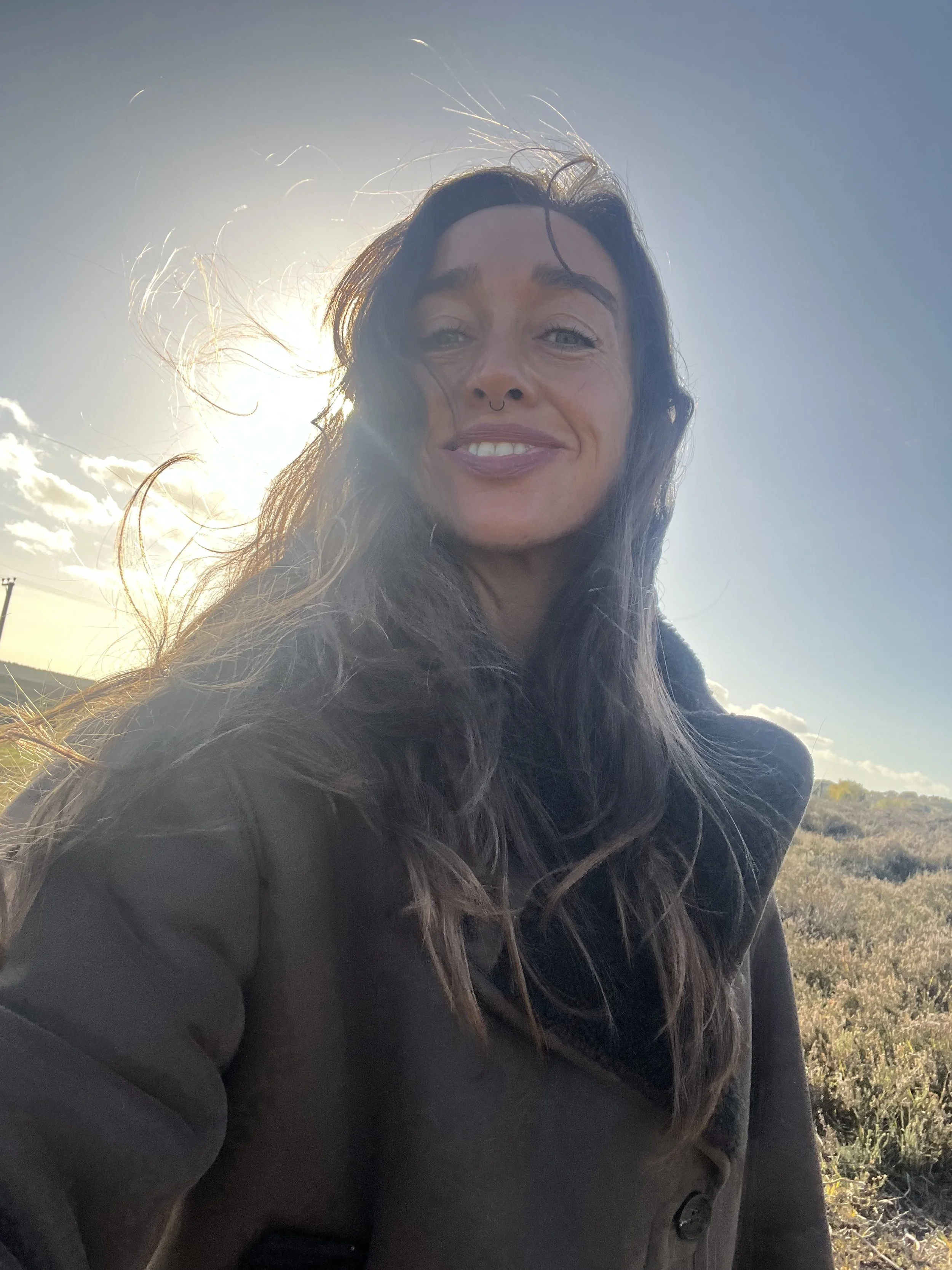 Close-up of a woman with long dark hair, wearing a dark coat, smiling outdoors with the sun behind her and a clear blue sky.