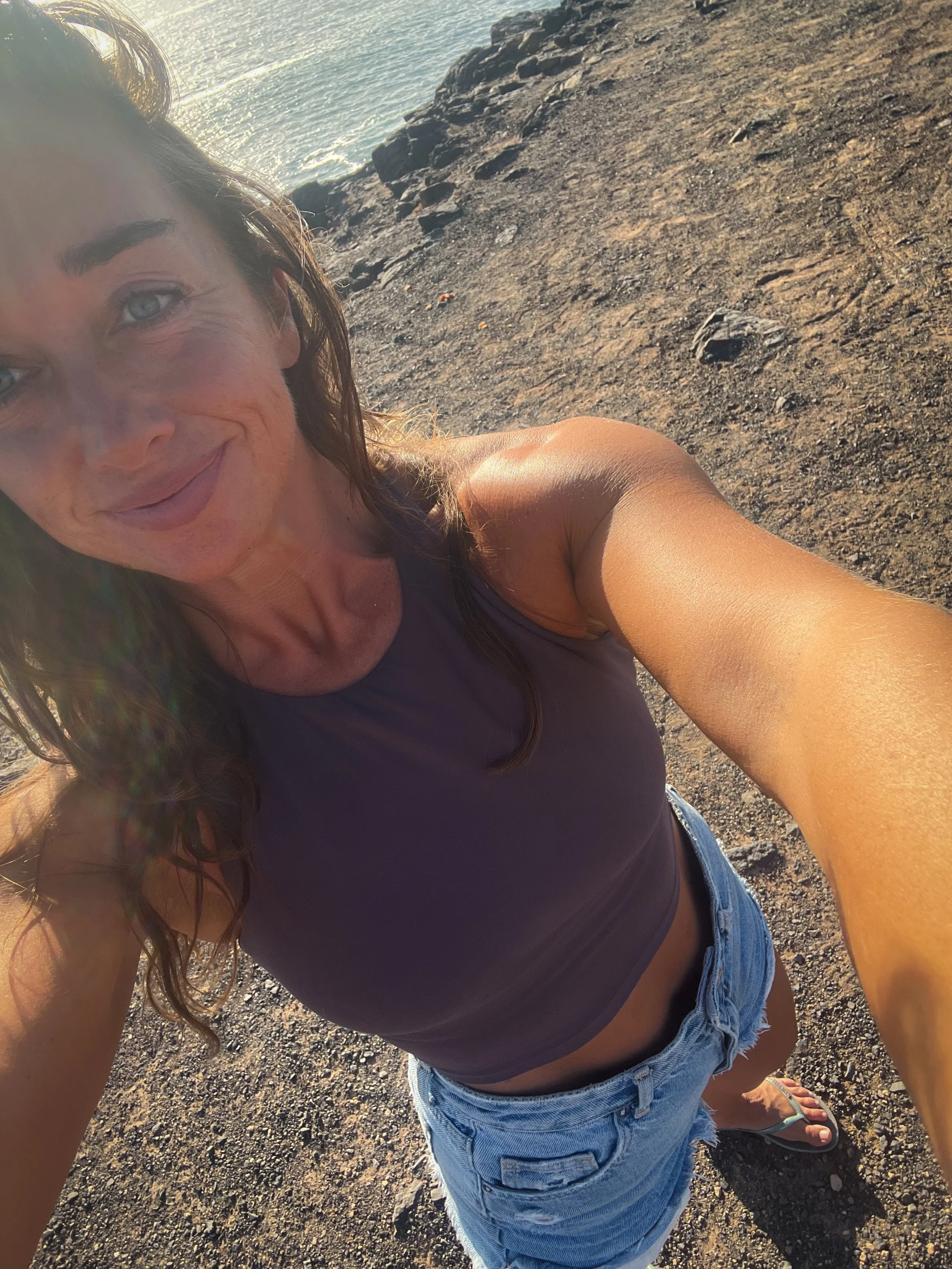 A woman taking a selfie on a rocky beach during sunset, wearing a dark tank top, denim shorts, and flip-flops.