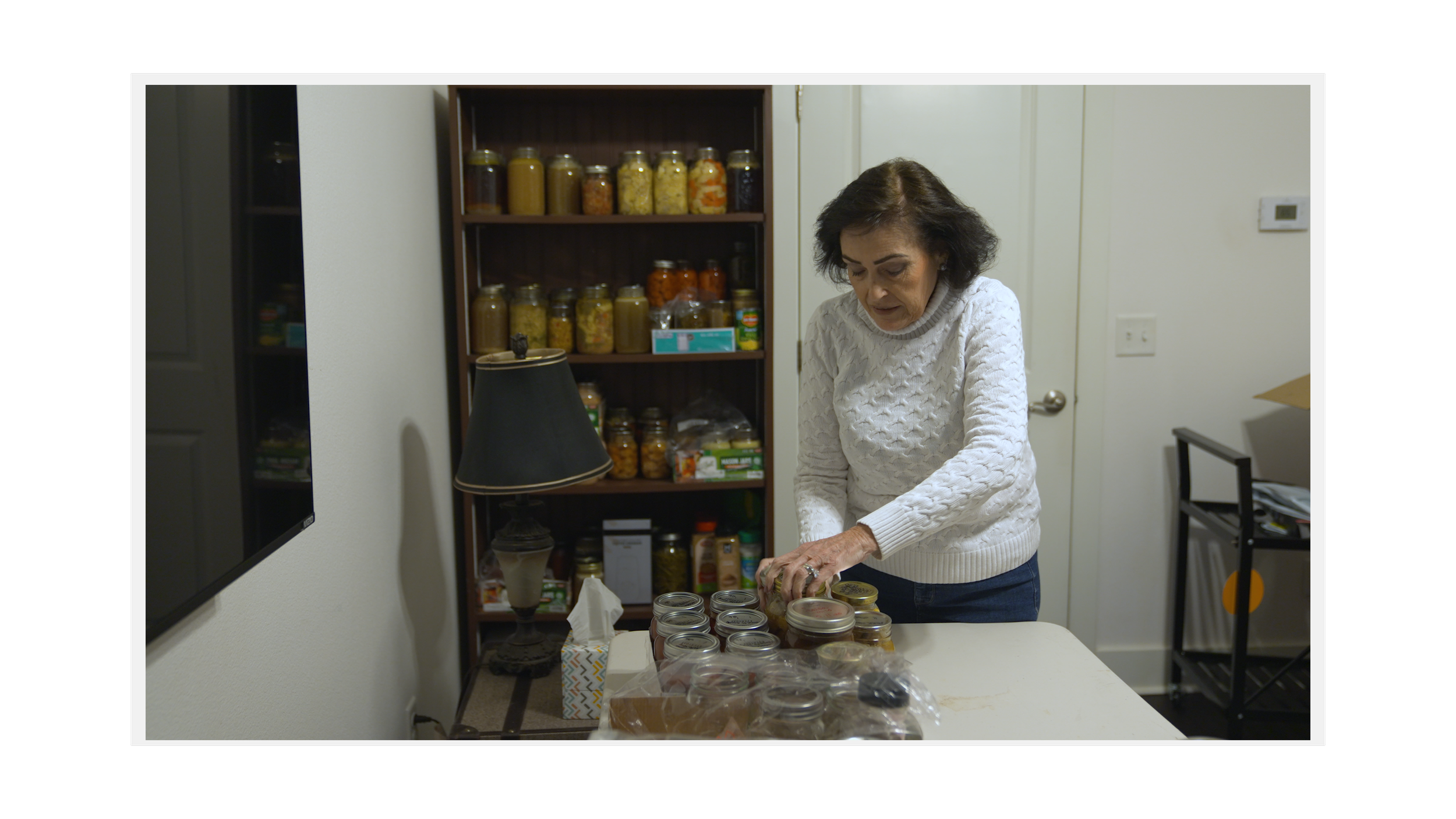 Woman organizing jars on a table in a pantry with shelves of canned goods in the background.