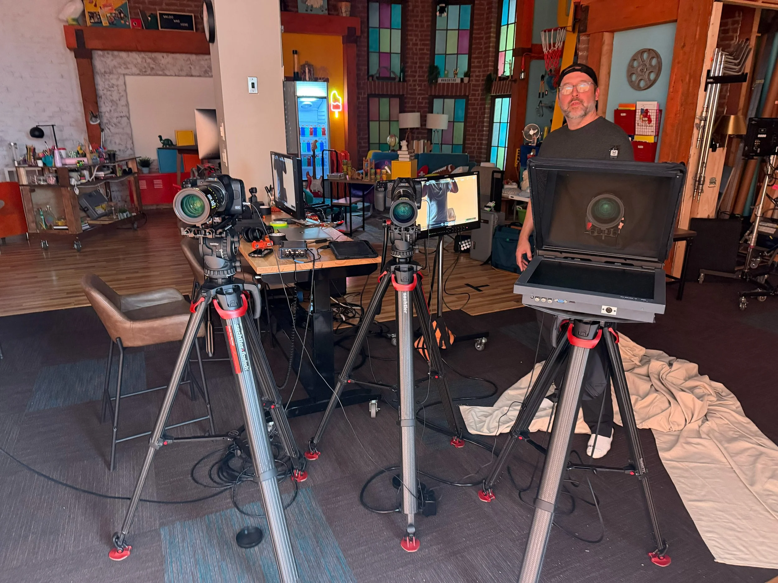 Photo of a recording or film set inside a brightly decorated, colorful studio or workspace with a man standing near cameras and monitors.