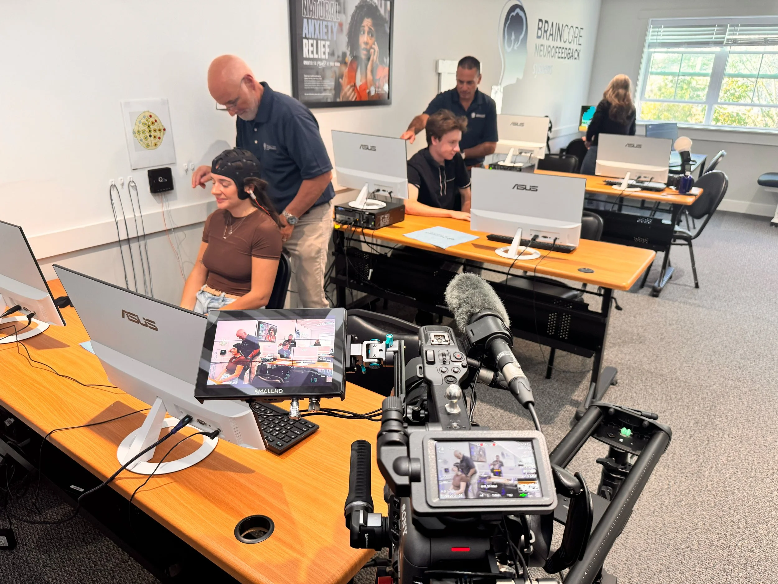 People working in a computer lab, with cameras and filming equipment capturing the scene. The room has multiple desks with monitors, and the walls have educational posters about mental health and neurofeedback.