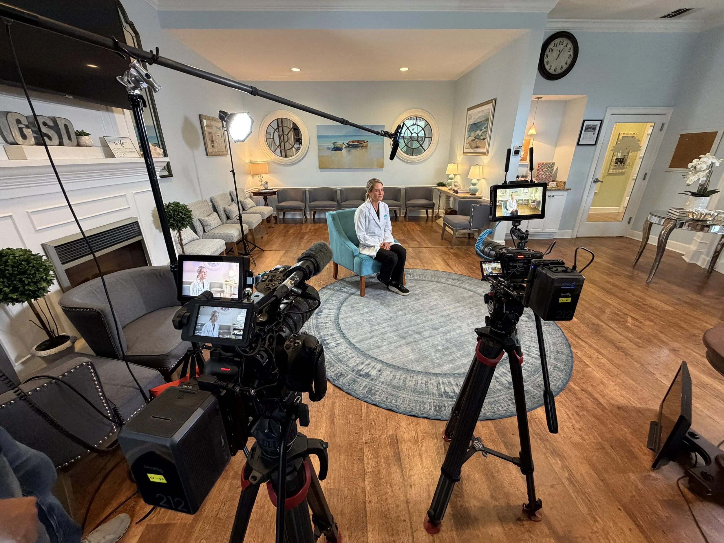 A woman in a white lab coat sitting on a teal chair in a well-lit living room with blue walls, surrounded by cameras and lights, during an interview or filming session.