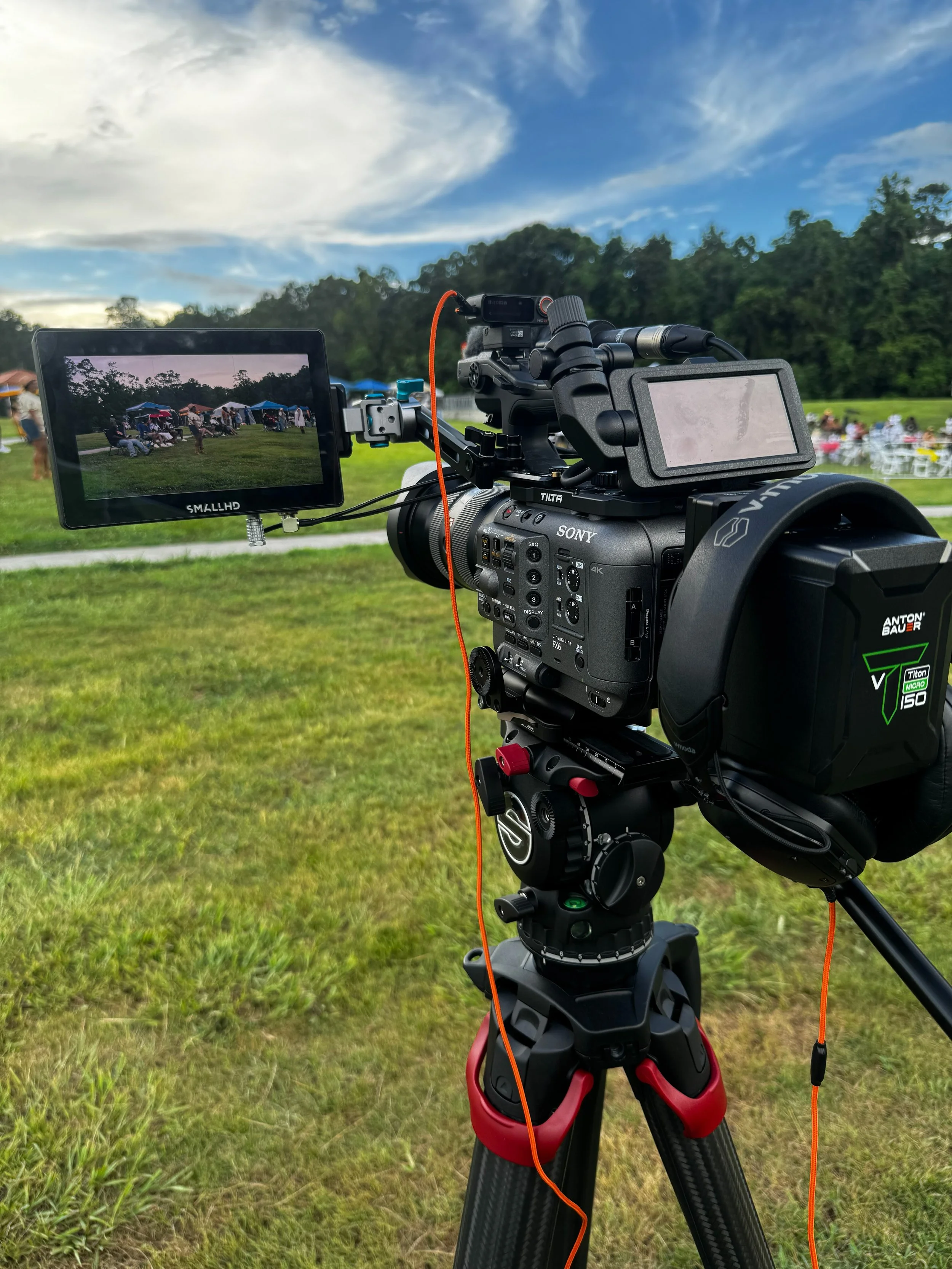 A professional video camera mounted on a tripod captures footage of an outdoor event. The camera has a monitor showing the scene of several people gathered on a grassy field with tents and chairs, under a sky with scattered clouds.