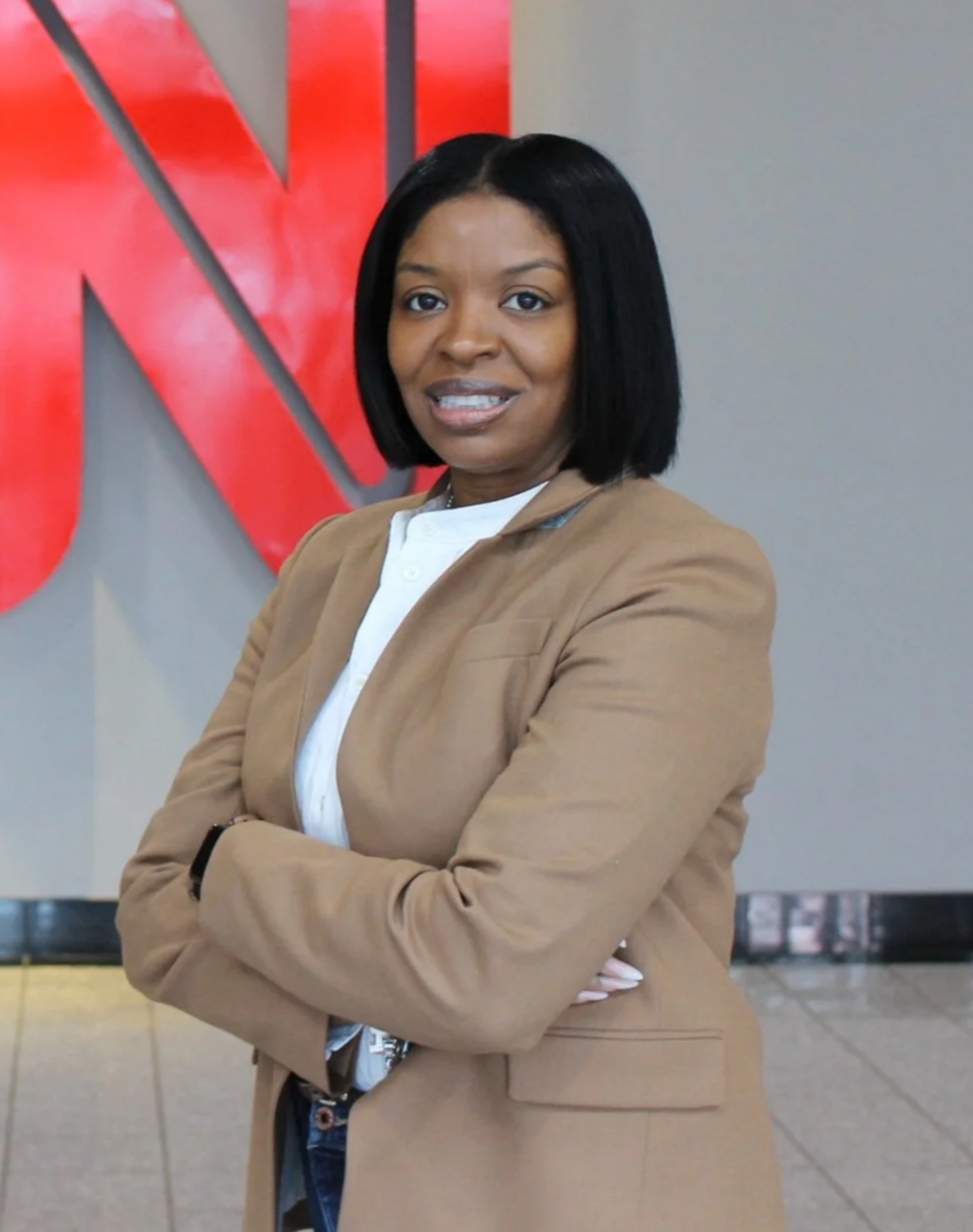 A woman with shoulder-length black hair, wearing a beige blazer and white shirt, standing with arms crossed in front of a gray wall with a red logo, looking at the camera.
