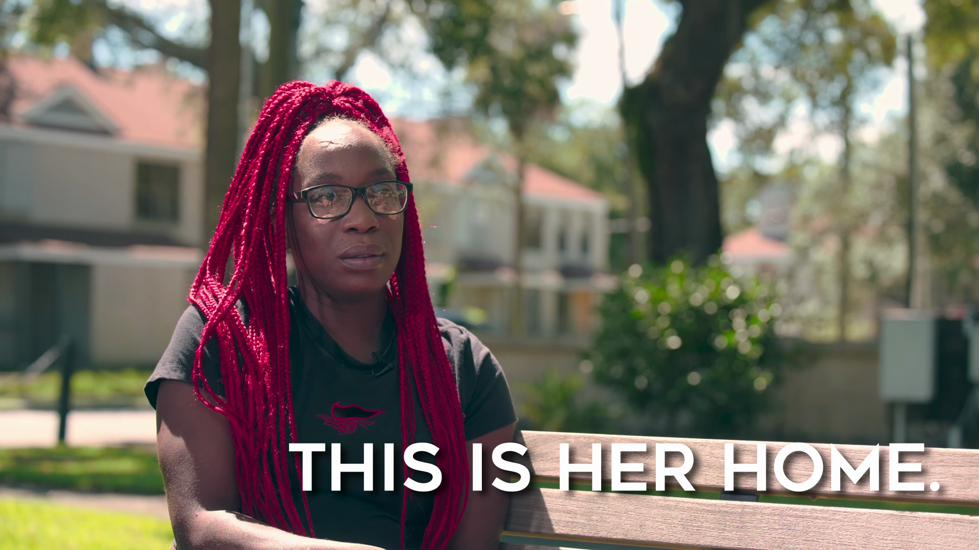 A woman with red braided hair and glasses sitting on a park bench outdoors, with houses and trees in the background. The text overlay says "This is her home."