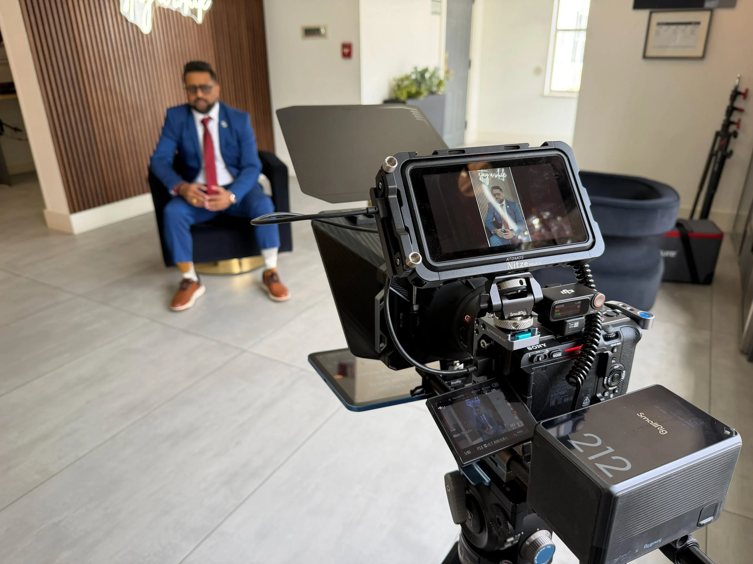 A man in a blue suit and red tie is sitting in a chair, holding a phone, during a video shoot. Camera equipment is pointed at him, capturing his image on a monitor.