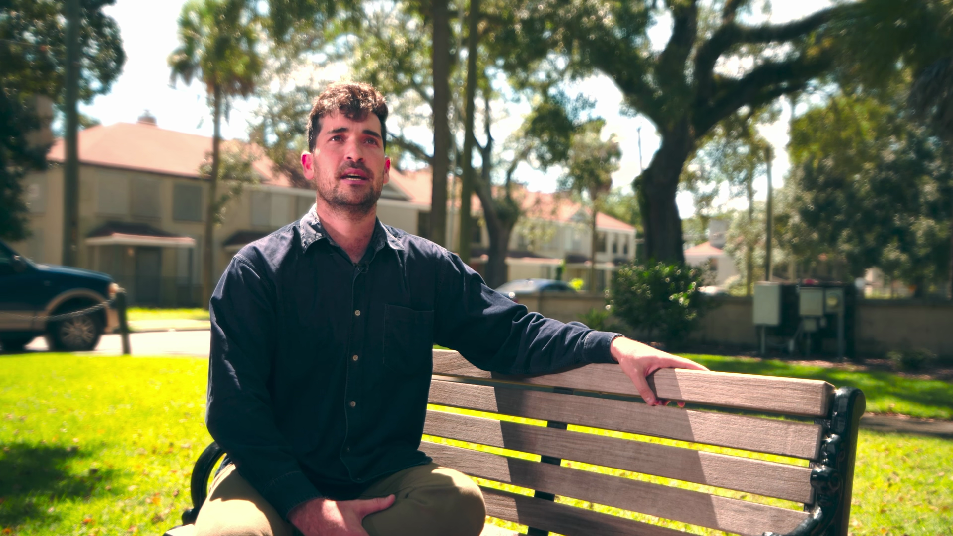 A young man with dark hair and facial hair sitting on a park bench, wearing a dark blue button-up shirt and khaki pants, with trees, houses, and parked cars in the background on a sunny day.
