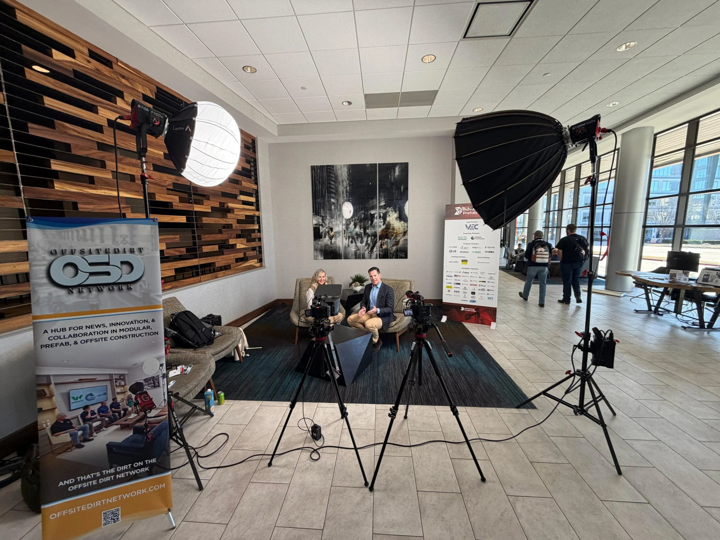 An indoor event space with studio lighting equipment, two people seated on a couch, and a promotional banner for Offsite Dirt Network. The space has large windows, a decorative wood wall, and a painting on the wall.