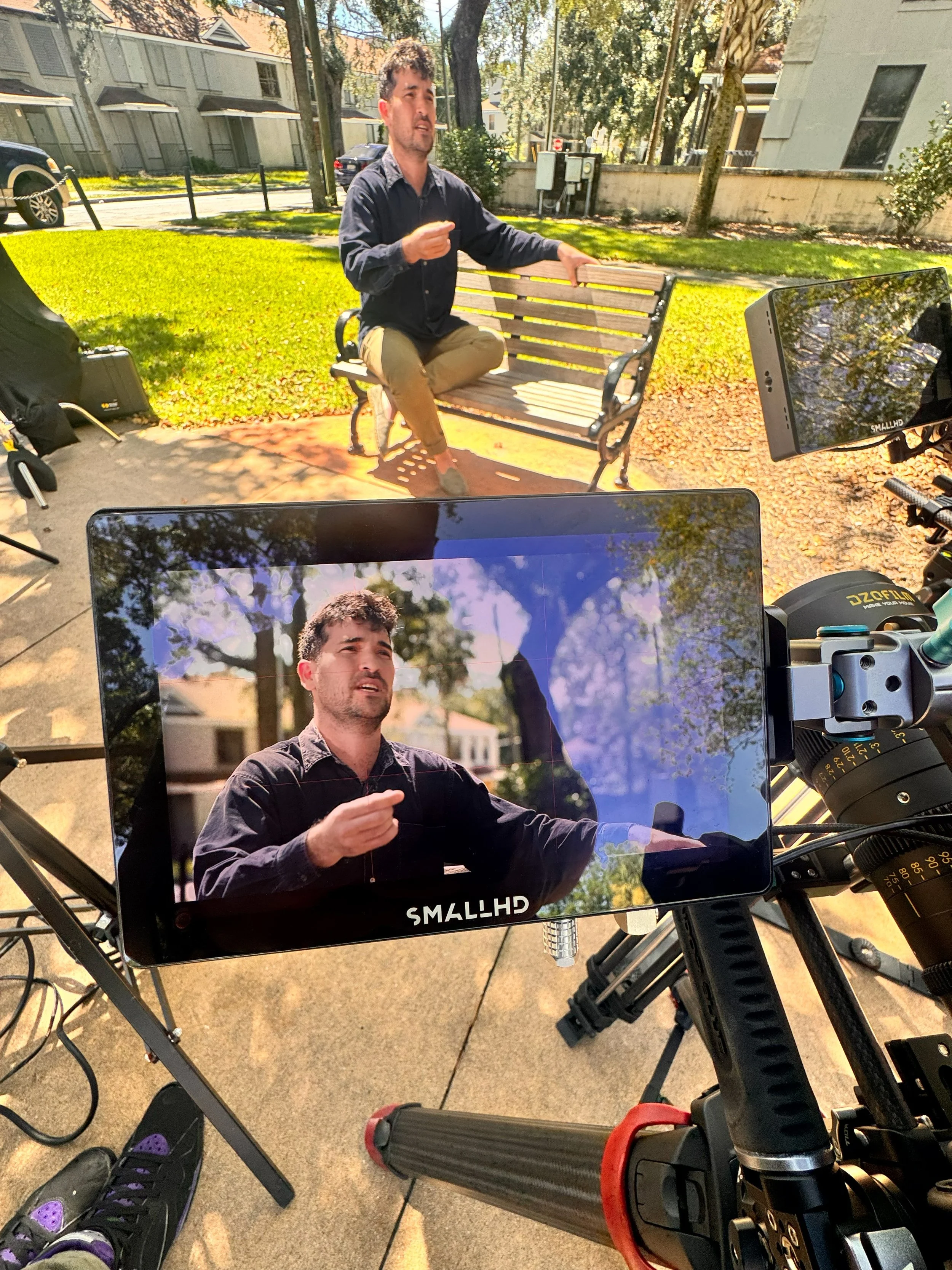 A man with dark hair and a beard, wearing a dark shirt and khaki pants, sits on a wooden park bench outdoors. He appears to be speaking and gesturing with his right hand. There is a smartphone mounted on a tripod in front of him, reflecting his image