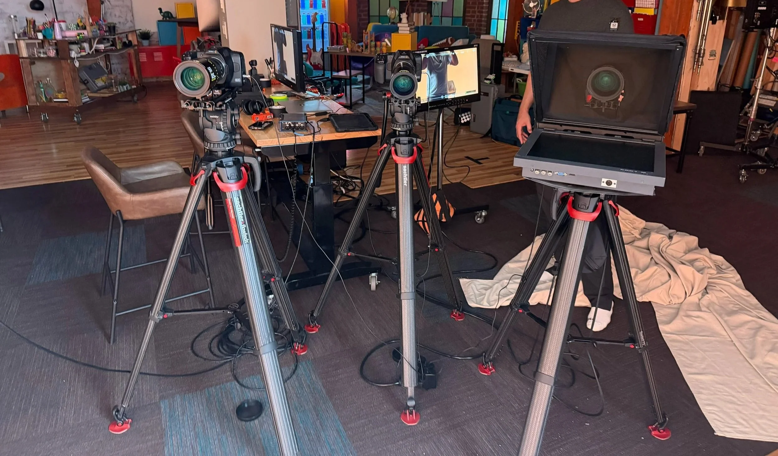A room set up for filming with professional camera equipment on tripods directed at a television monitor and a computer screen. There are chairs, a blanket on the floor, and various office supplies and decorations in the background.