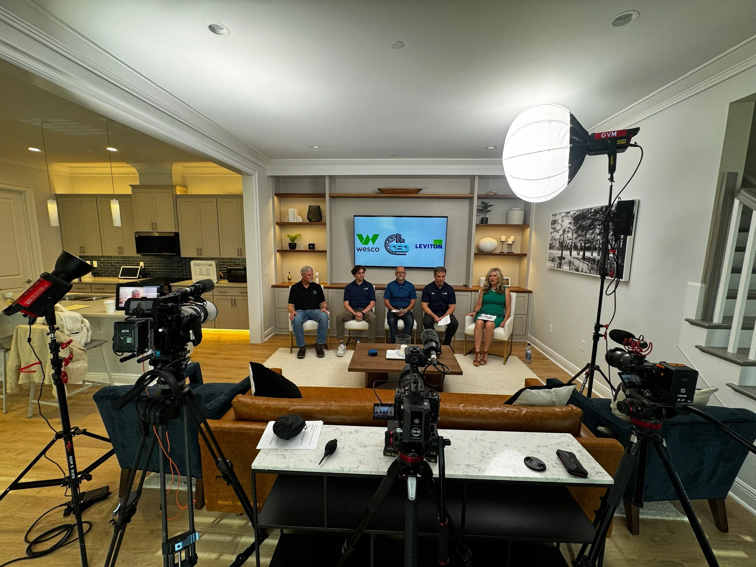 A panel of five speakers, four men and one woman, sitting in a room with a large screen behind them displaying logos for Wesco, Leviton, and another company. The room is set up for a video recording or live stream with professional camera equipment a