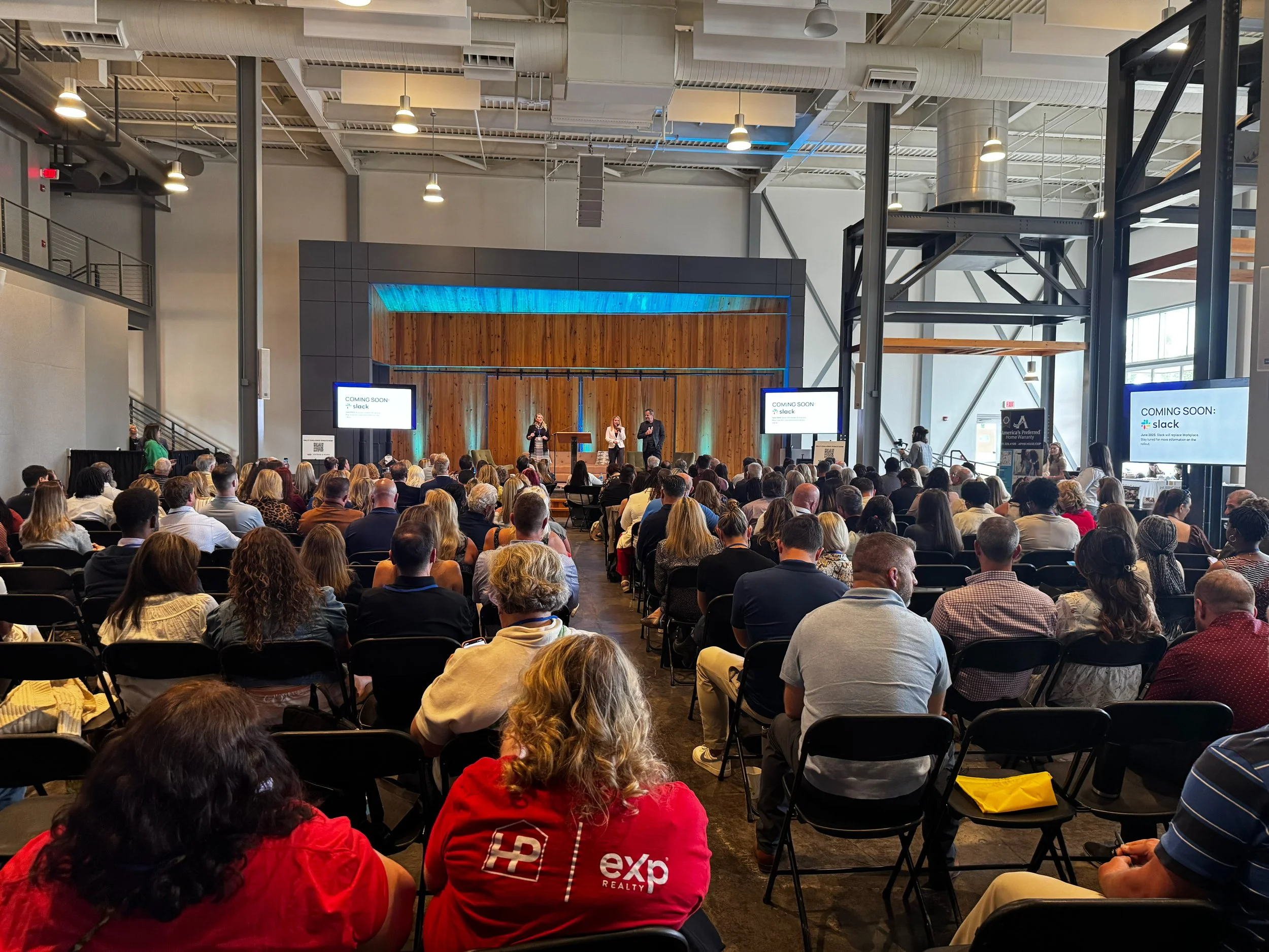 Large indoor event with a stage and an audience seated in chairs. Three speakers are on stage, with two screens displaying 'COMING SOON: Slack.' The venue has high ceilings, exposed ductwork, and industrial design elements.