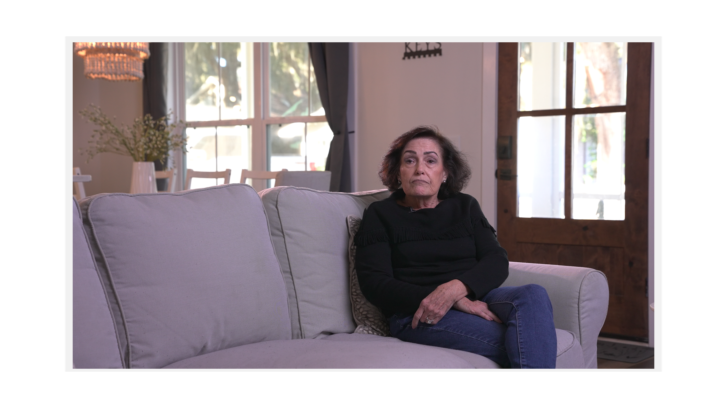 An older woman with dark hair sits on a light-colored sofa in a living room with large windows and wooden door. She is wearing a black top and blue jeans, with a neutral expression.