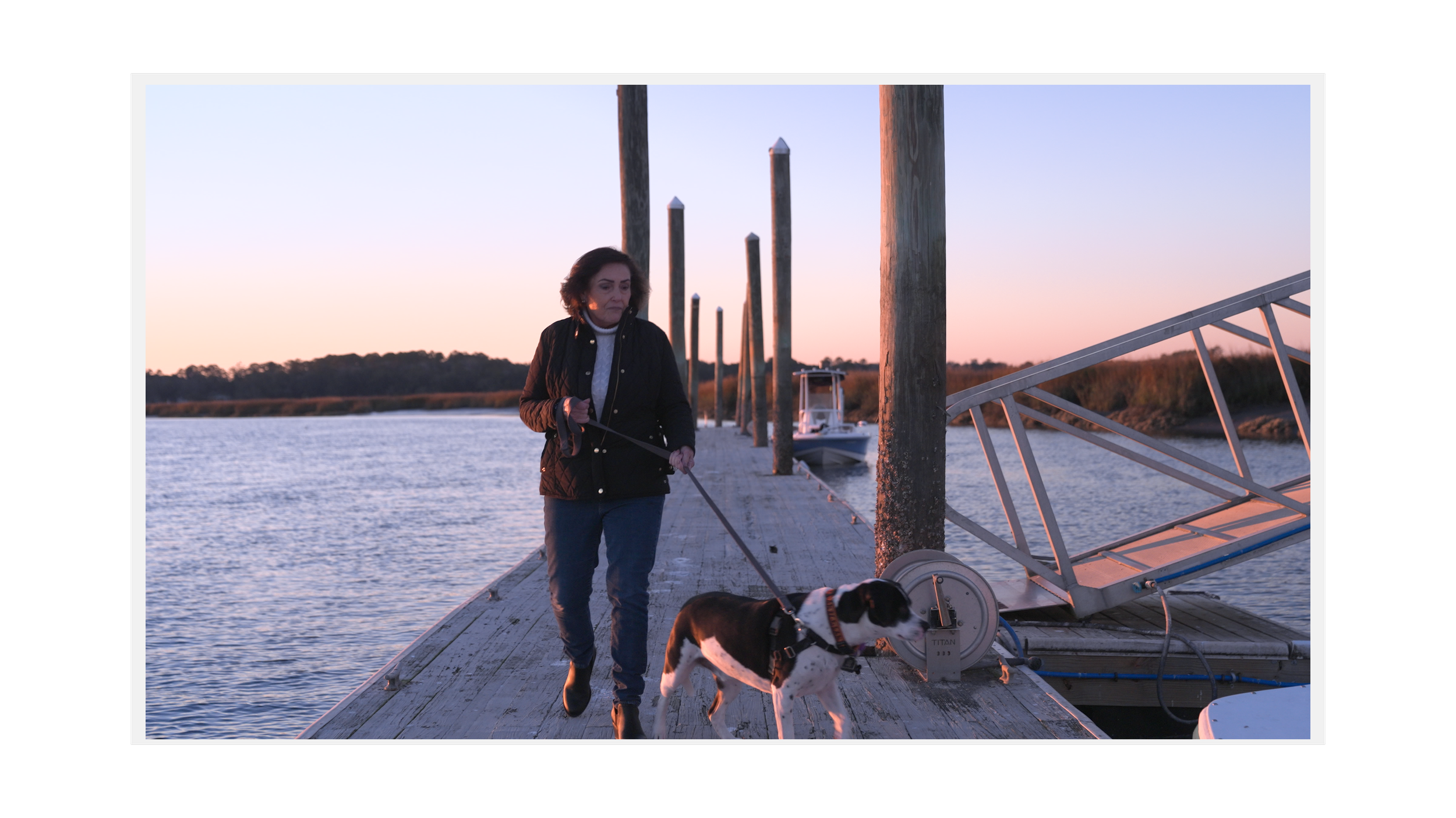 Woman walking her dog on a dock by the water at sunset.