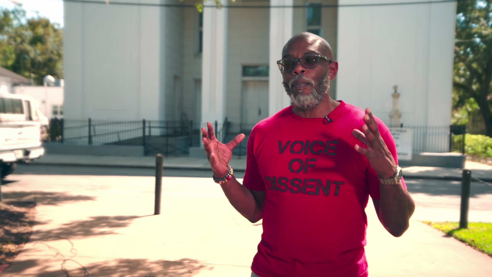 A man with glasses, a gray beard, and a red shirt with the words "Voice of Dissent" printed on it, speaking outdoors in front of a white building.